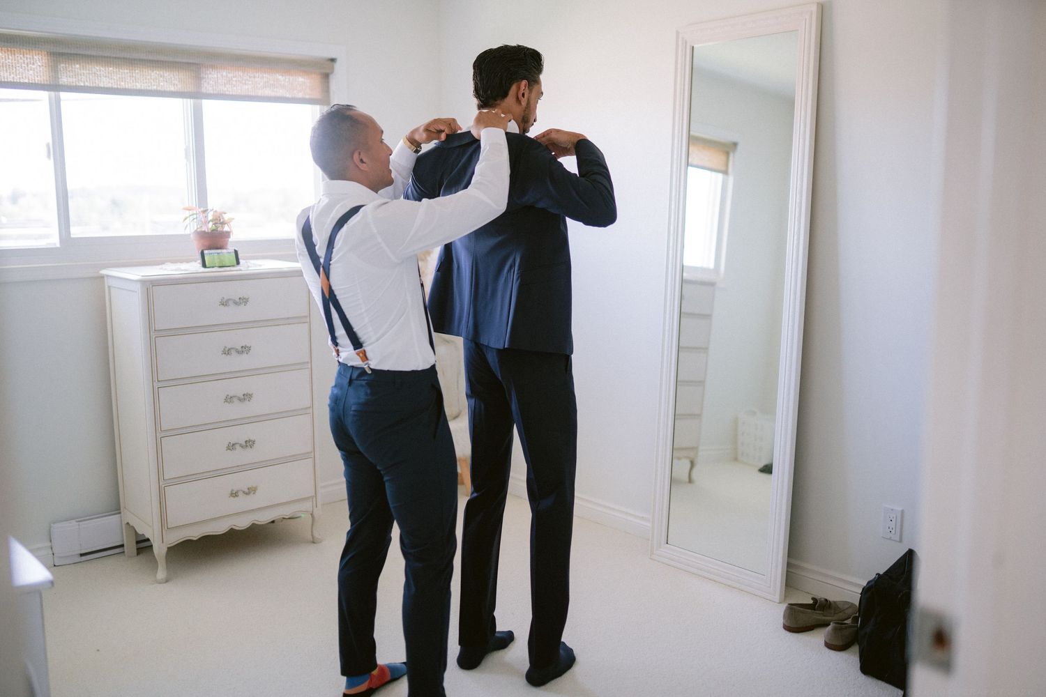 Father helping groom put on suit jacket in bedroom mirror during wedding day preparations.