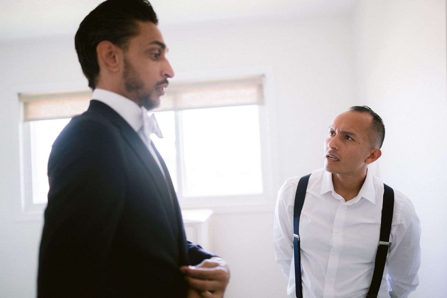 Groom in a navy suit shares a conversation with a groomsman wearing suspenders before the wedding.
