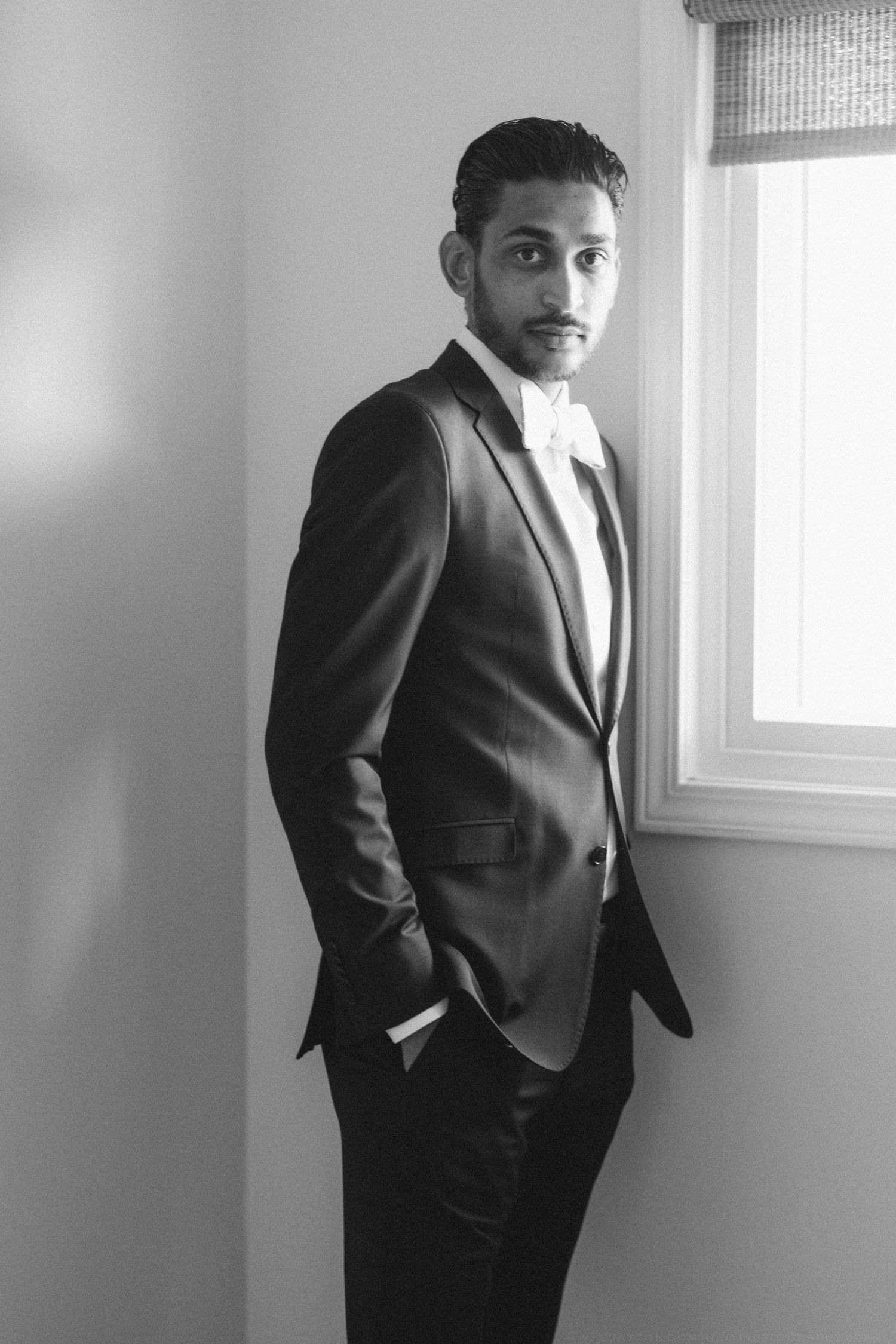 Handsome groom in black tuxedo with bow tie standing by window in black and white portrait.