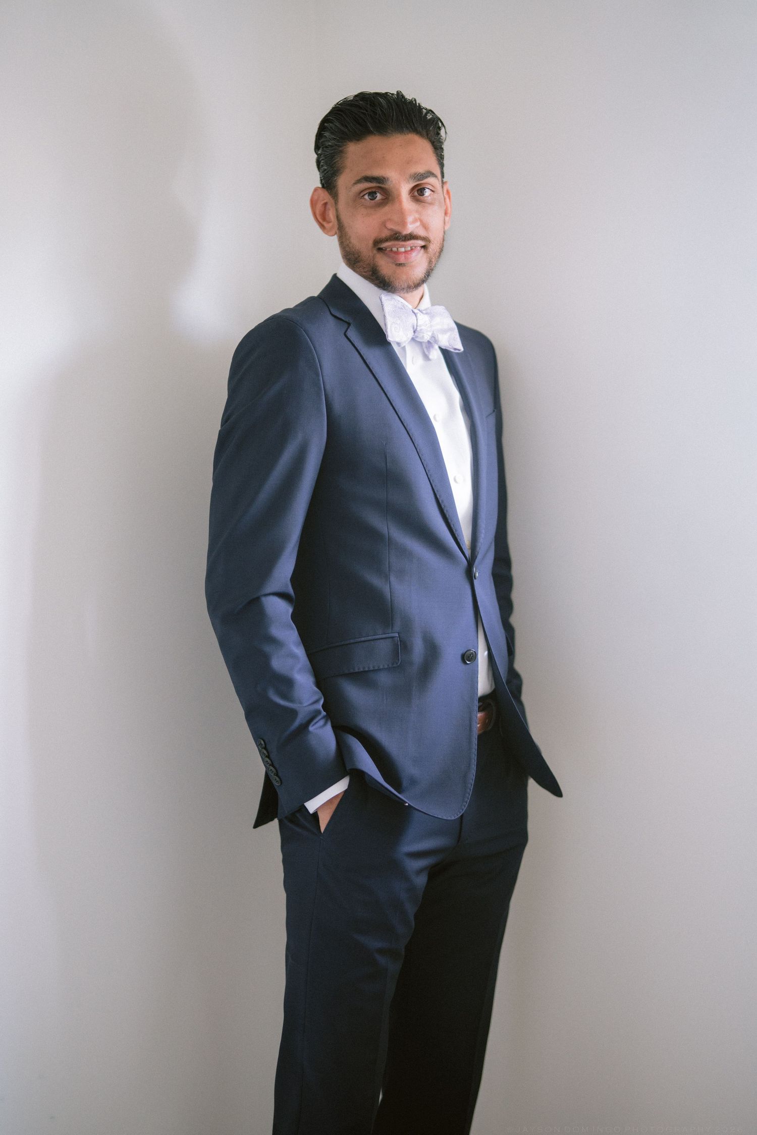 Groom in navy blue suit with white bow tie posing confidently against light gray background.