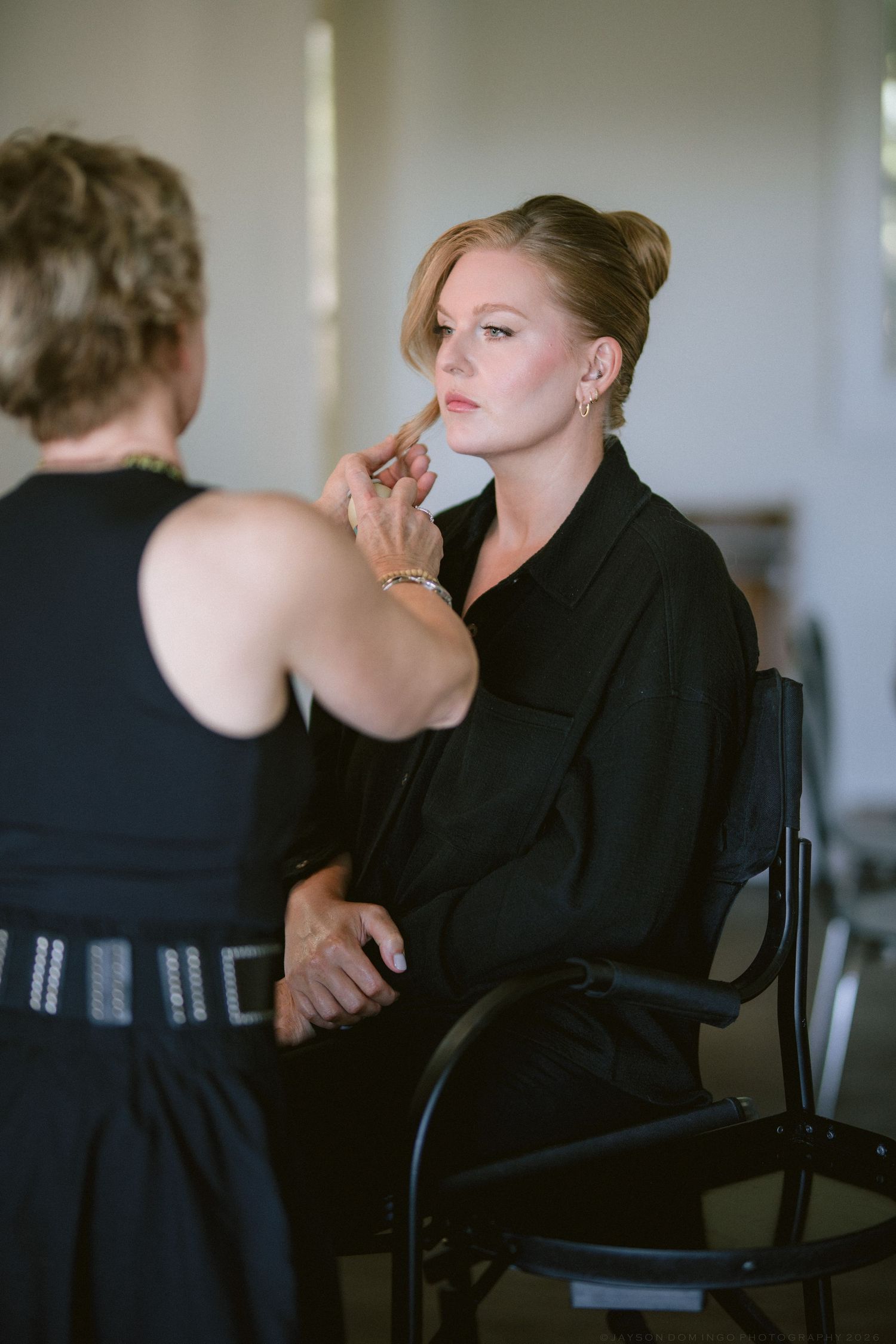 Makeup artist applying lipstick to blonde bride seated in chair during wedding preparation.