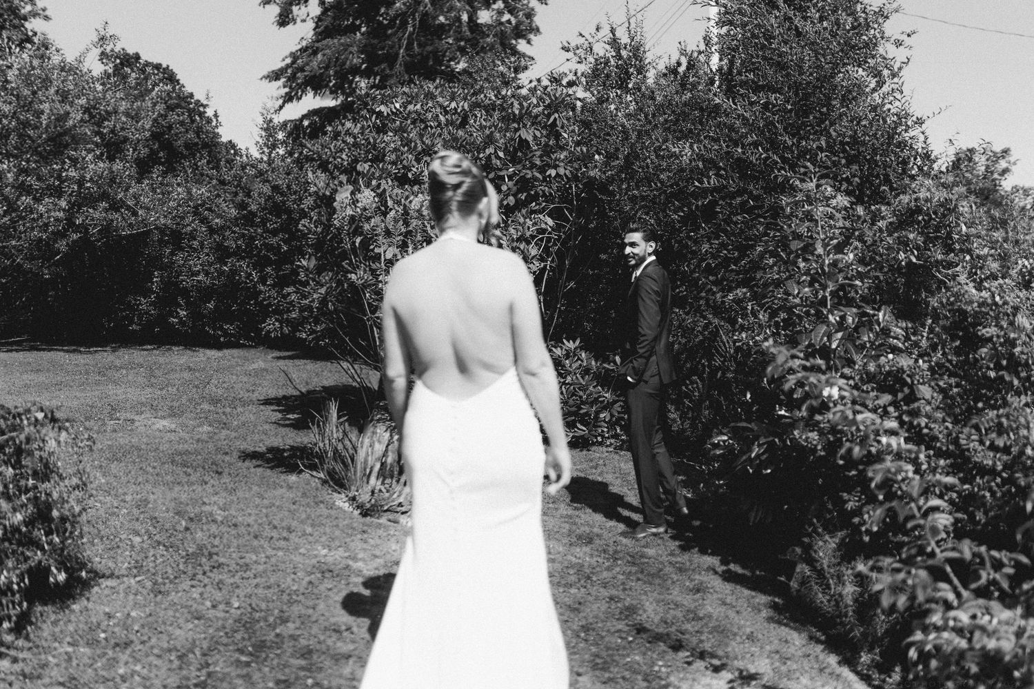 Black and white photo of bride in backless wedding gown walking toward groom in lush garden setting.