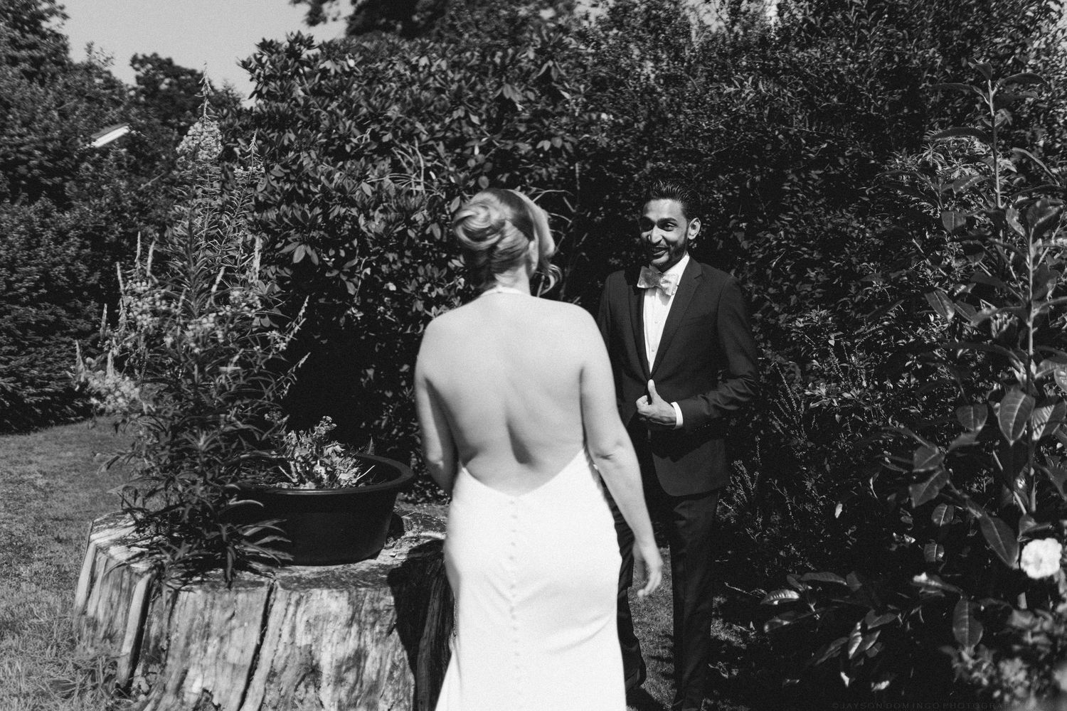 Black and white photo of groom smiling at bride during outdoor first look moment among lush greenery.