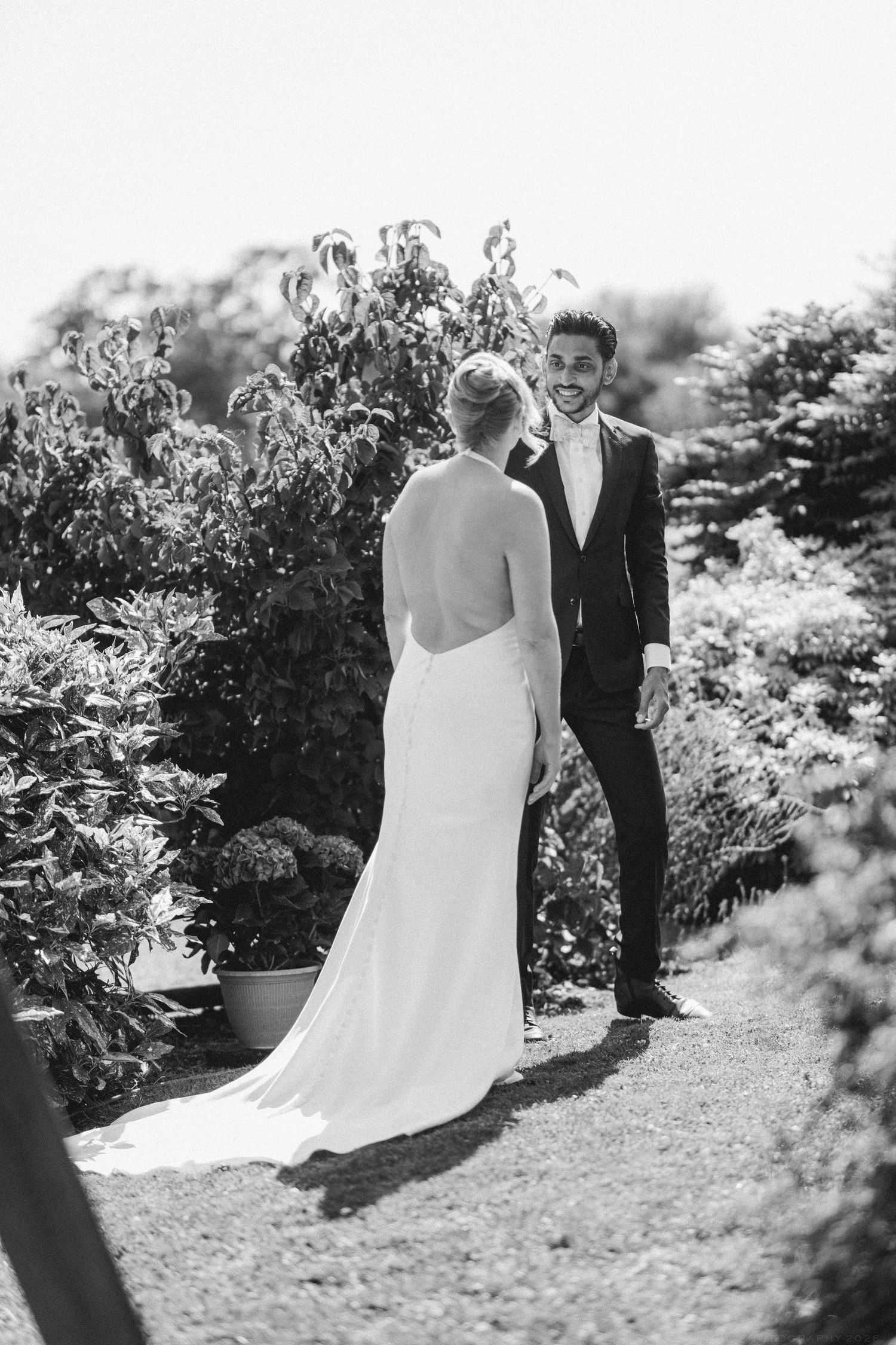 Black and white photo of a smiling groom in a suit seeing his bride in a backless wedding dress for the first time outdoors.