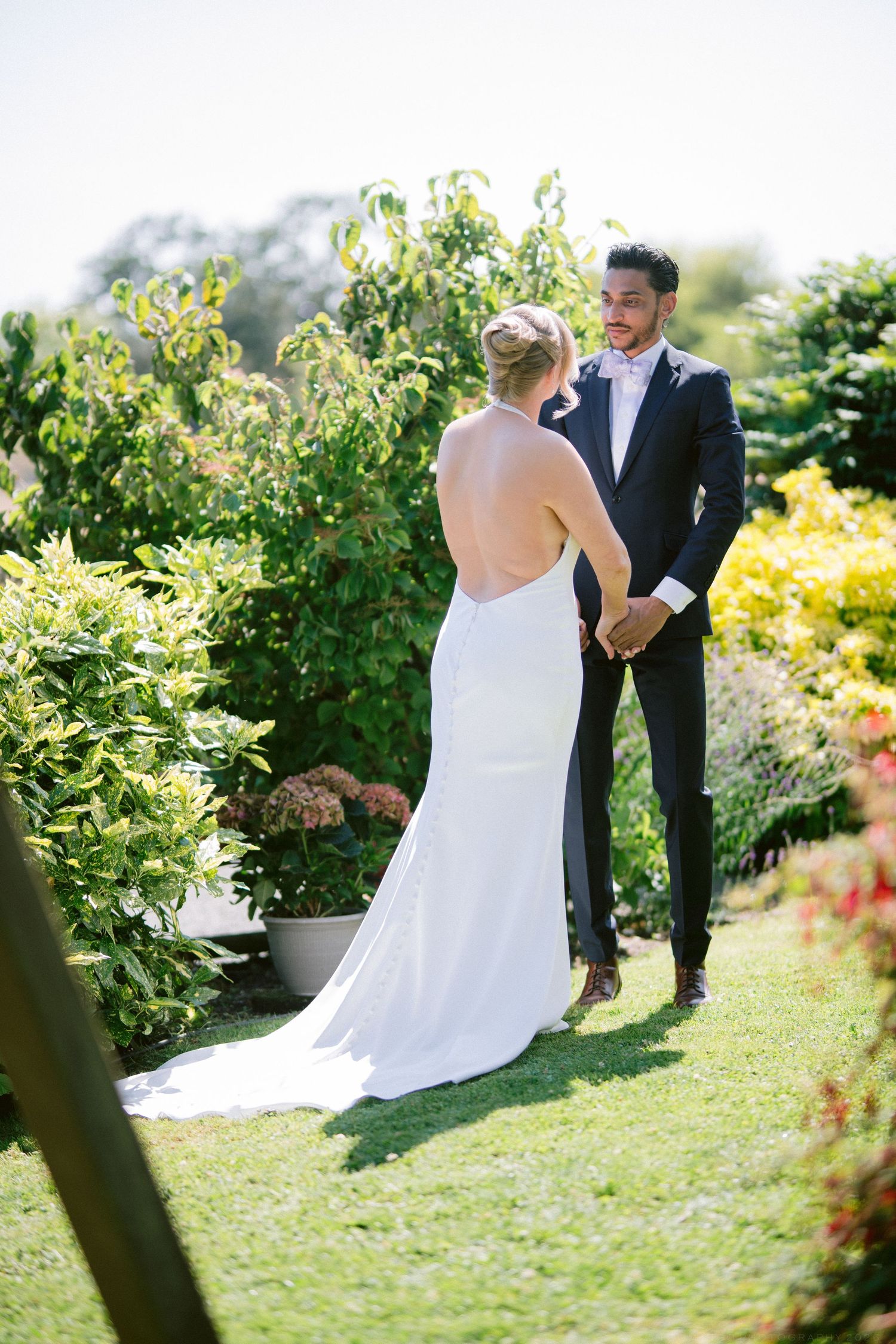 Bride and groom share first look moment in lush garden, holding hands on sunny wedding day.