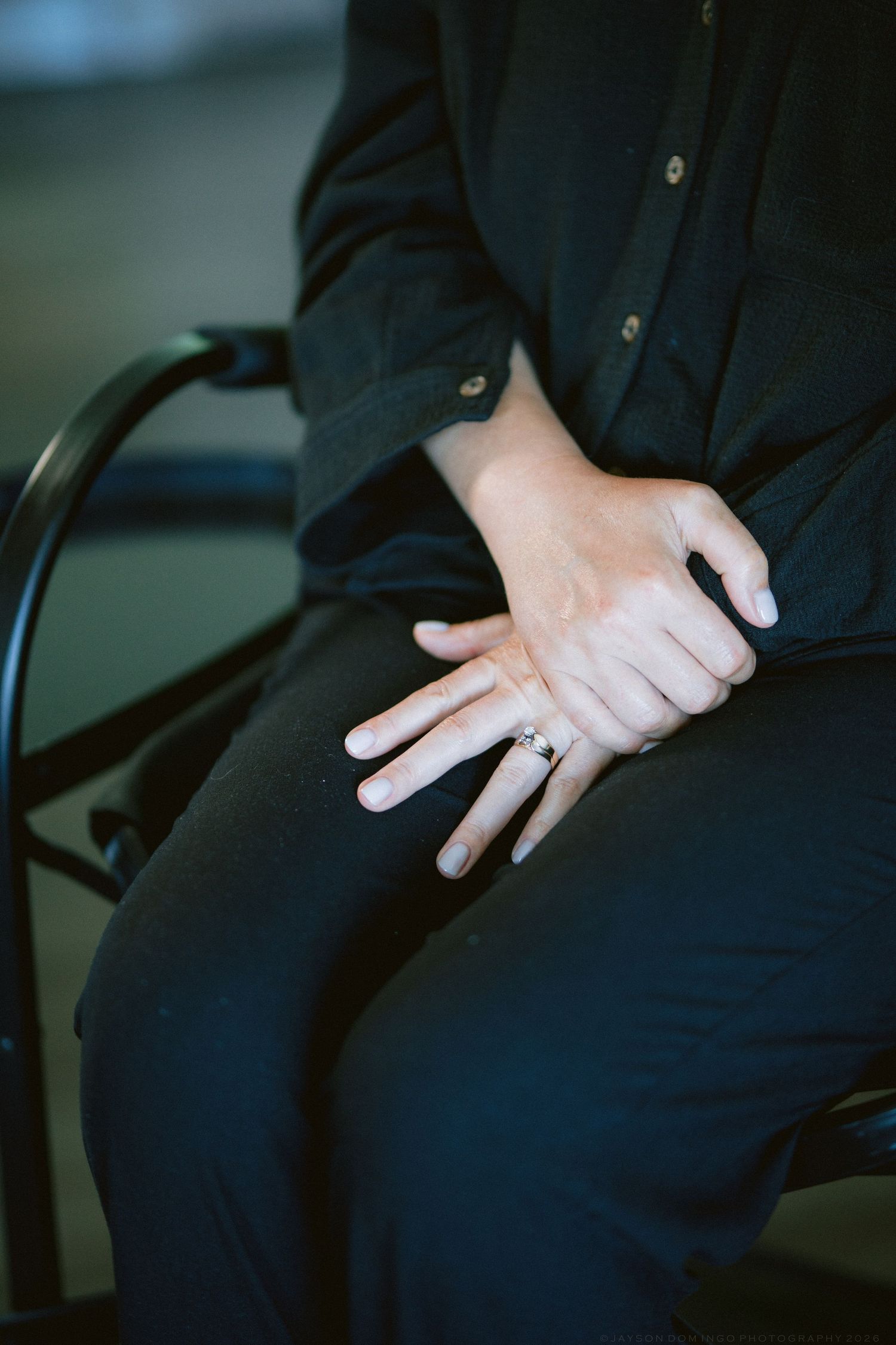 Close-up of bride's hands showing engagement ring while seated in black chair wearing all black outfit.
