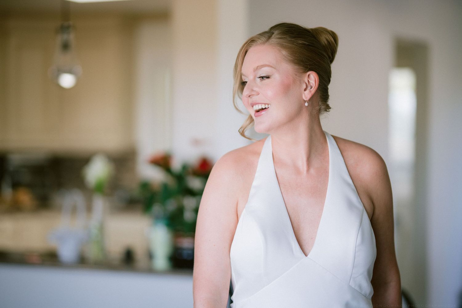 Radiant bride in white halter wedding dress smiles softly while getting ready indoors on wedding day.
