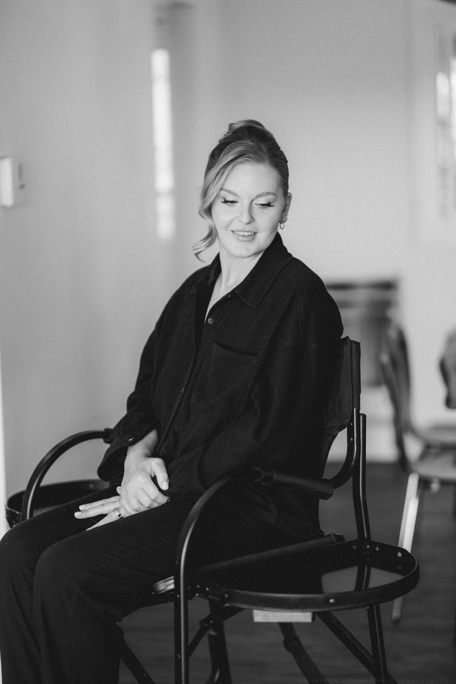 Black and white portrait of smiling blonde woman in dark jacket sitting relaxed on chair indoors.