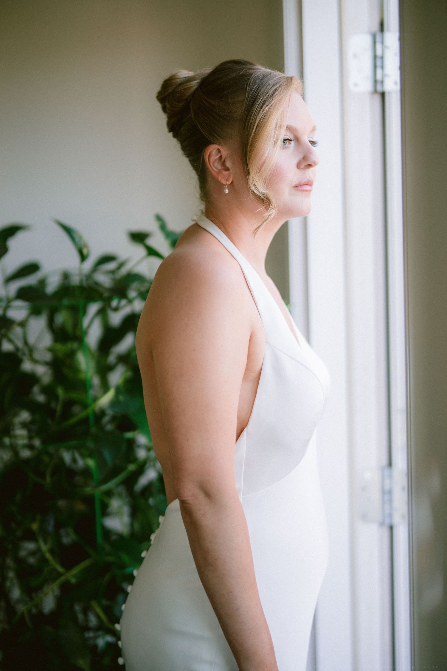 Bride in sleek white halter wedding dress gazing thoughtfully out a window surrounded by green plants.