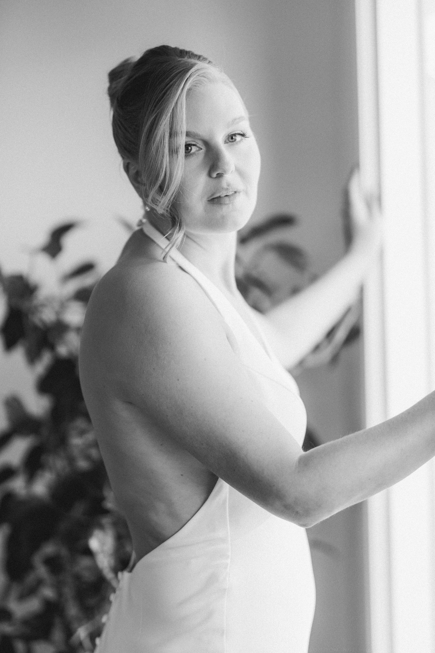 Black and white portrait of blonde bride in backless halter wedding gown standing by a bright window.