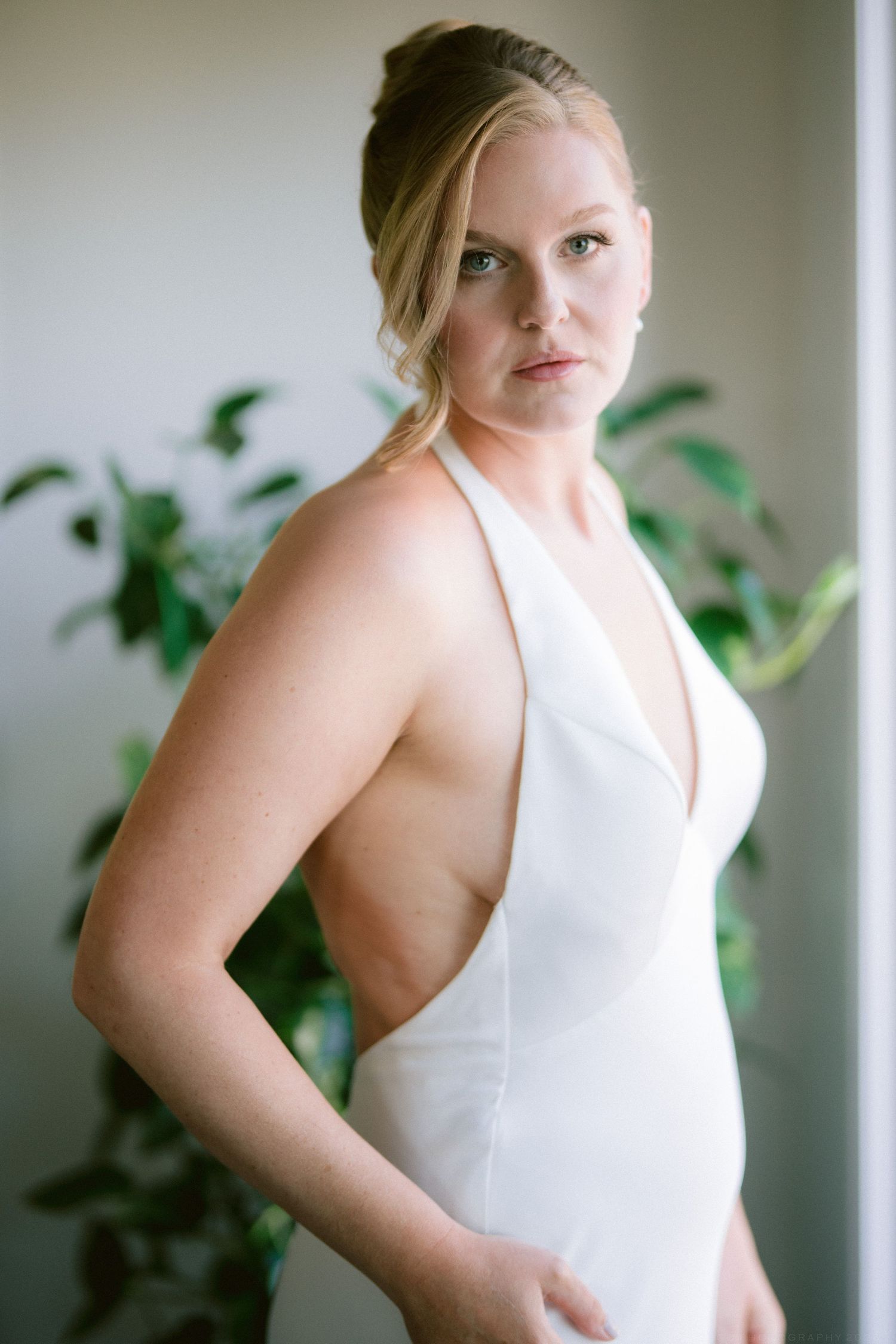 Blonde bride in a sleek white halter neck wedding dress posing elegantly by a window.