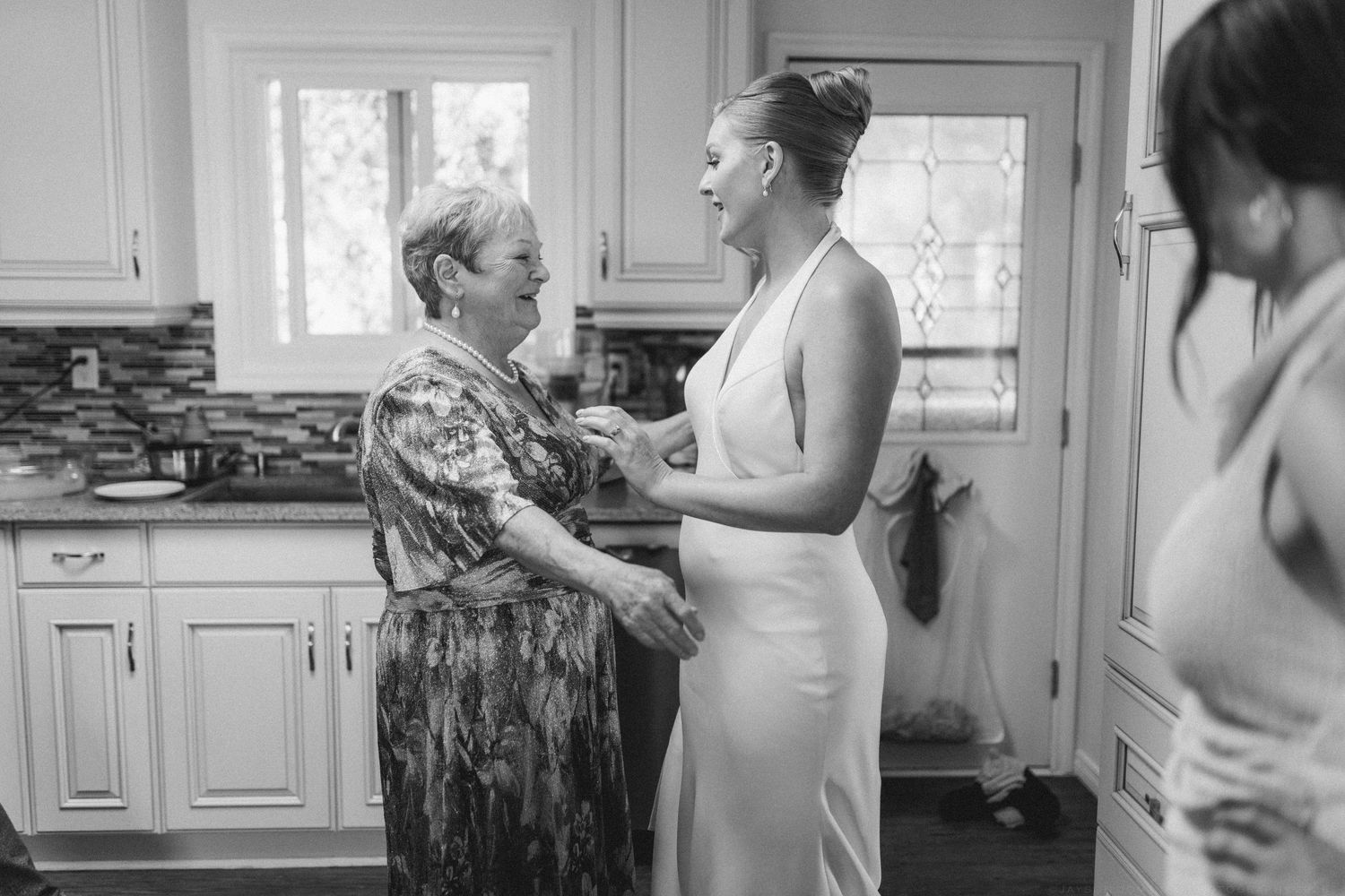 Bride in white gown laughing with older woman in kitchen during wedding preparations, black and white photo.