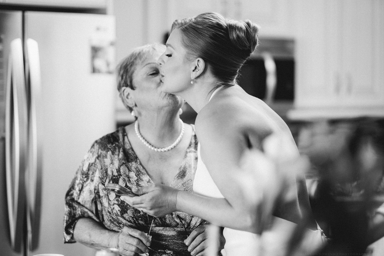 Black and white photo of bride sharing a tender kiss with older woman, likely mother, in kitchen.
