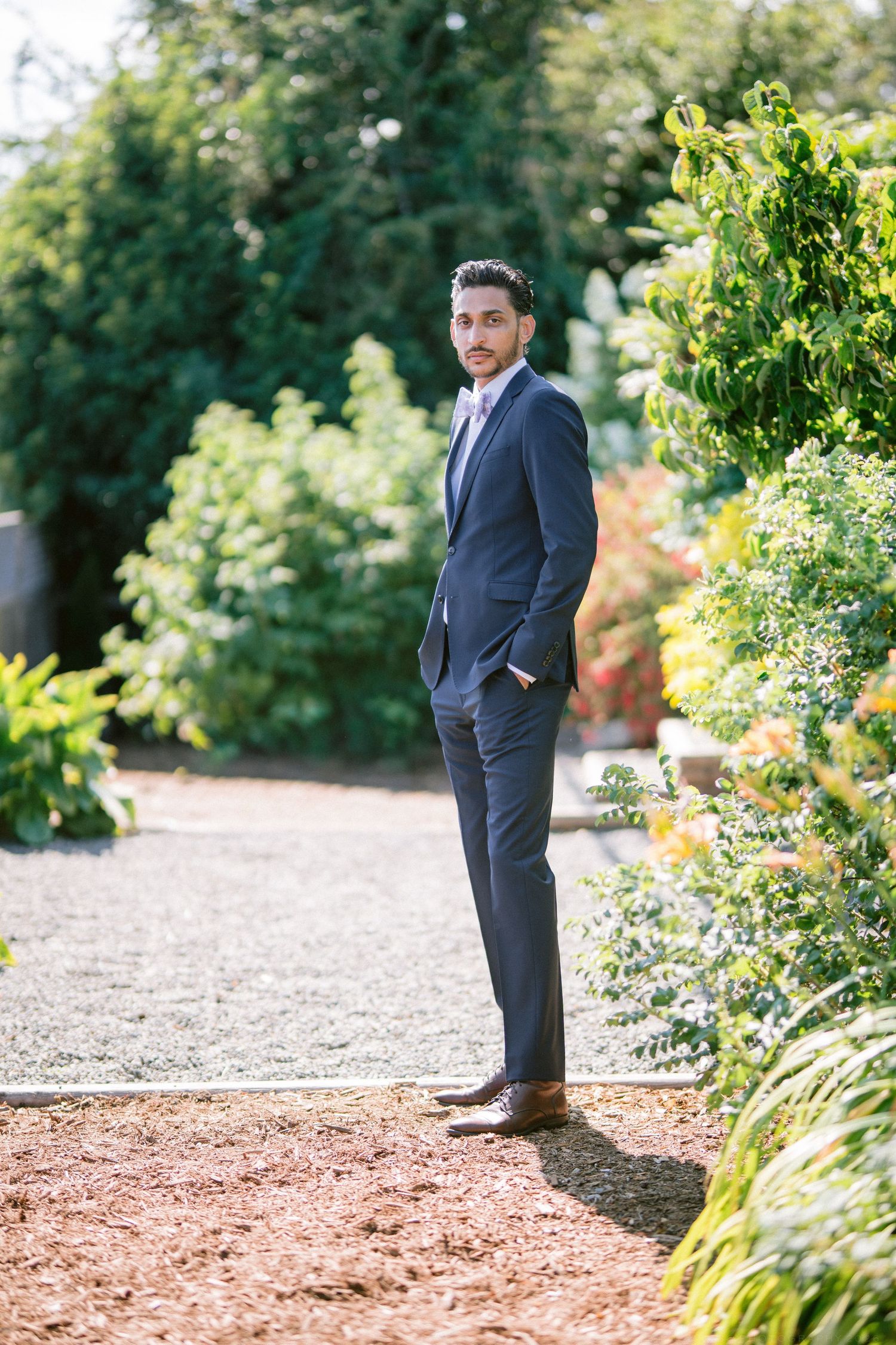 Dapper groom in navy suit standing on garden path with hands in pockets surrounded by lush greenery.