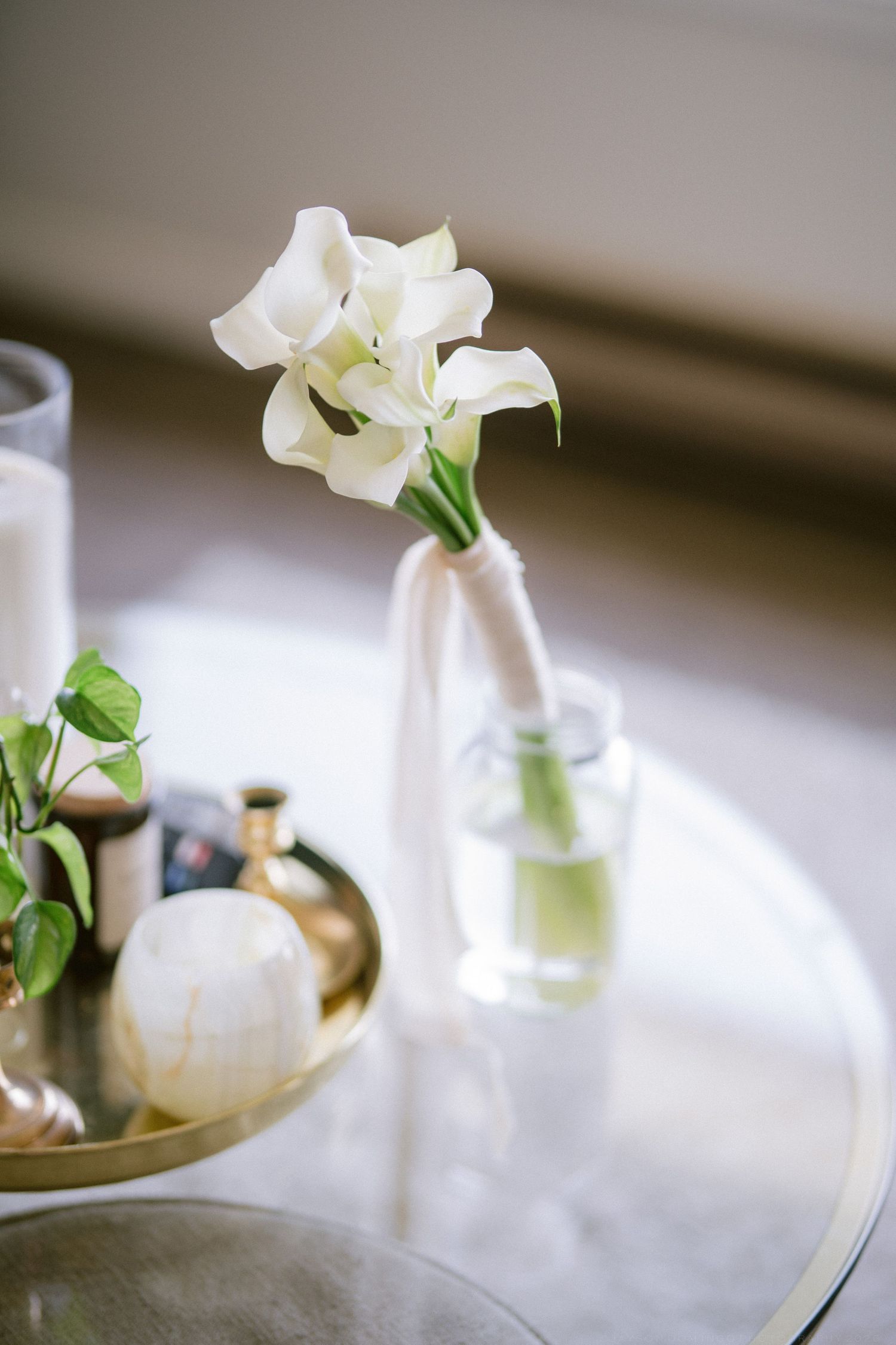 White calla lily bouquet in glass vase on marble table with gold decorative tray and candles at wedding reception.