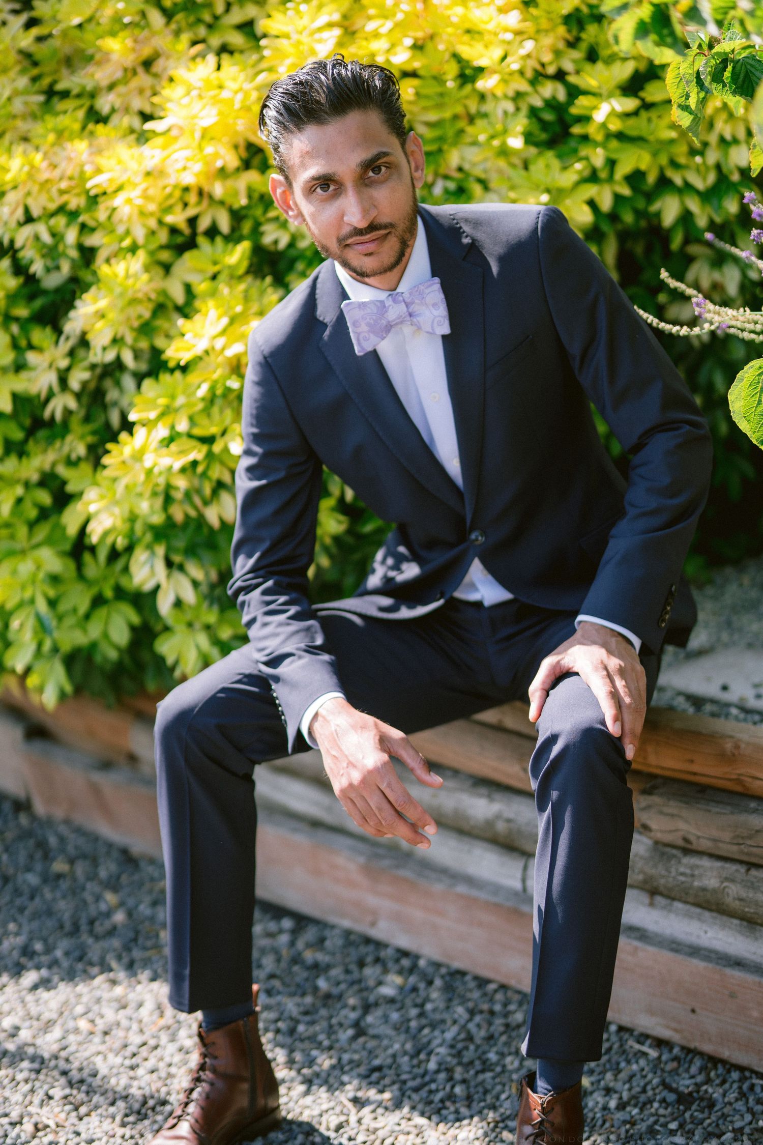 Groom in navy suit and bow tie sitting outdoors surrounded by lush green foliage.