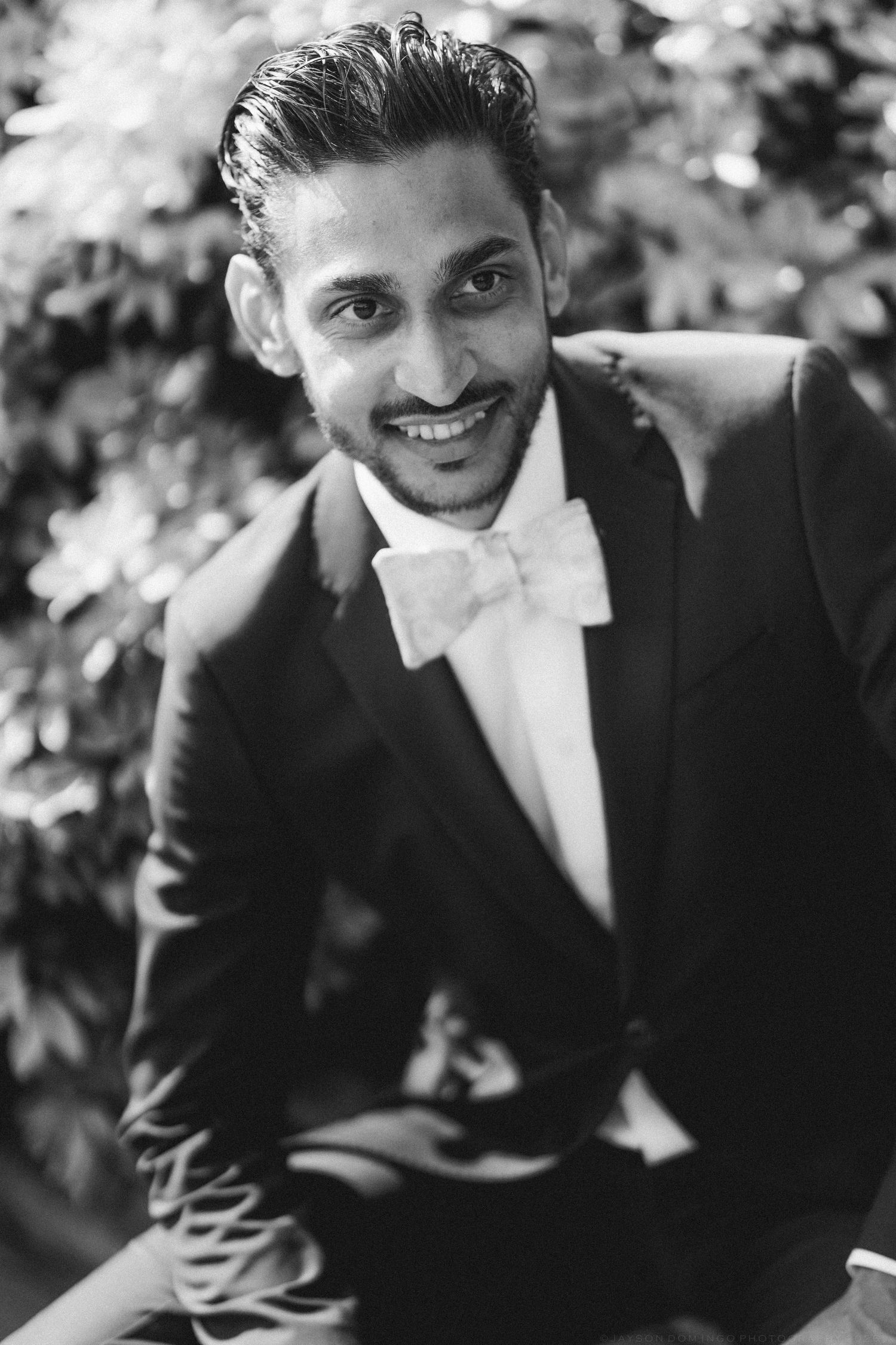 Smiling groom in black tuxedo with bow tie posing outdoors in black and white wedding portrait.