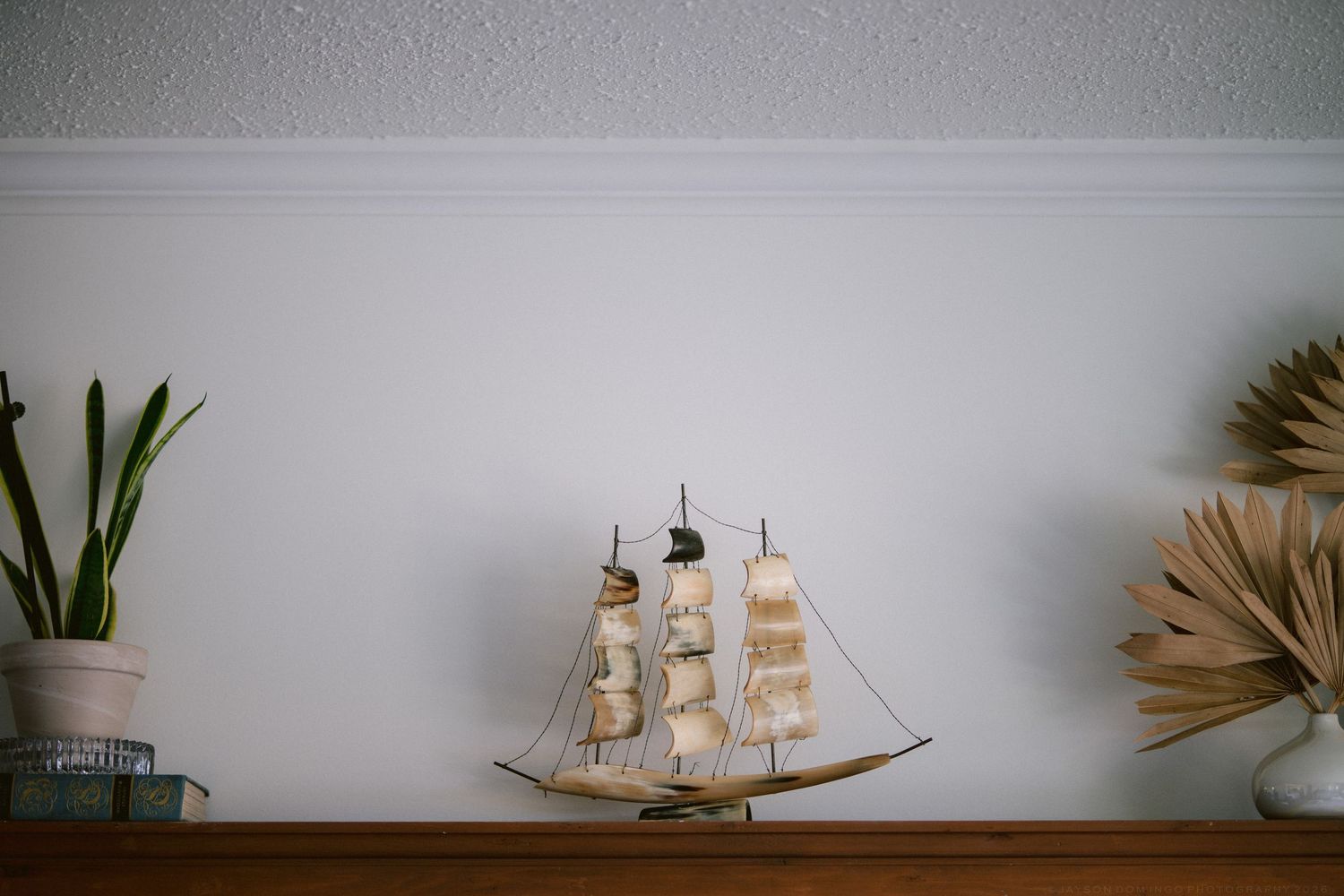 Decorative model sailboat displayed on wooden shelf with small plants and dried botanicals against white wall.