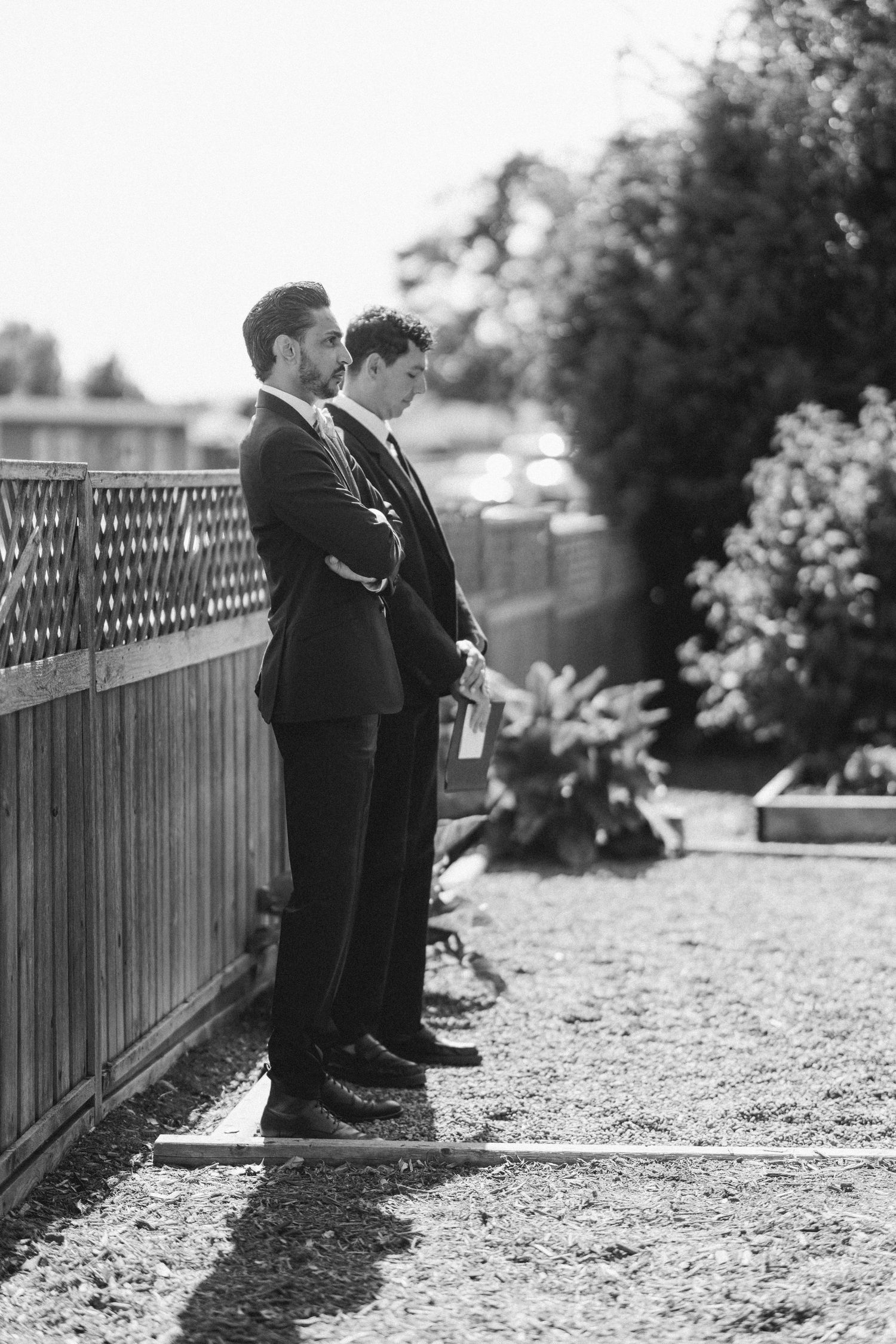 Two groomsmen in dark suits stand solemnly by a wooden fence in a sunlit garden before a wedding ceremony.