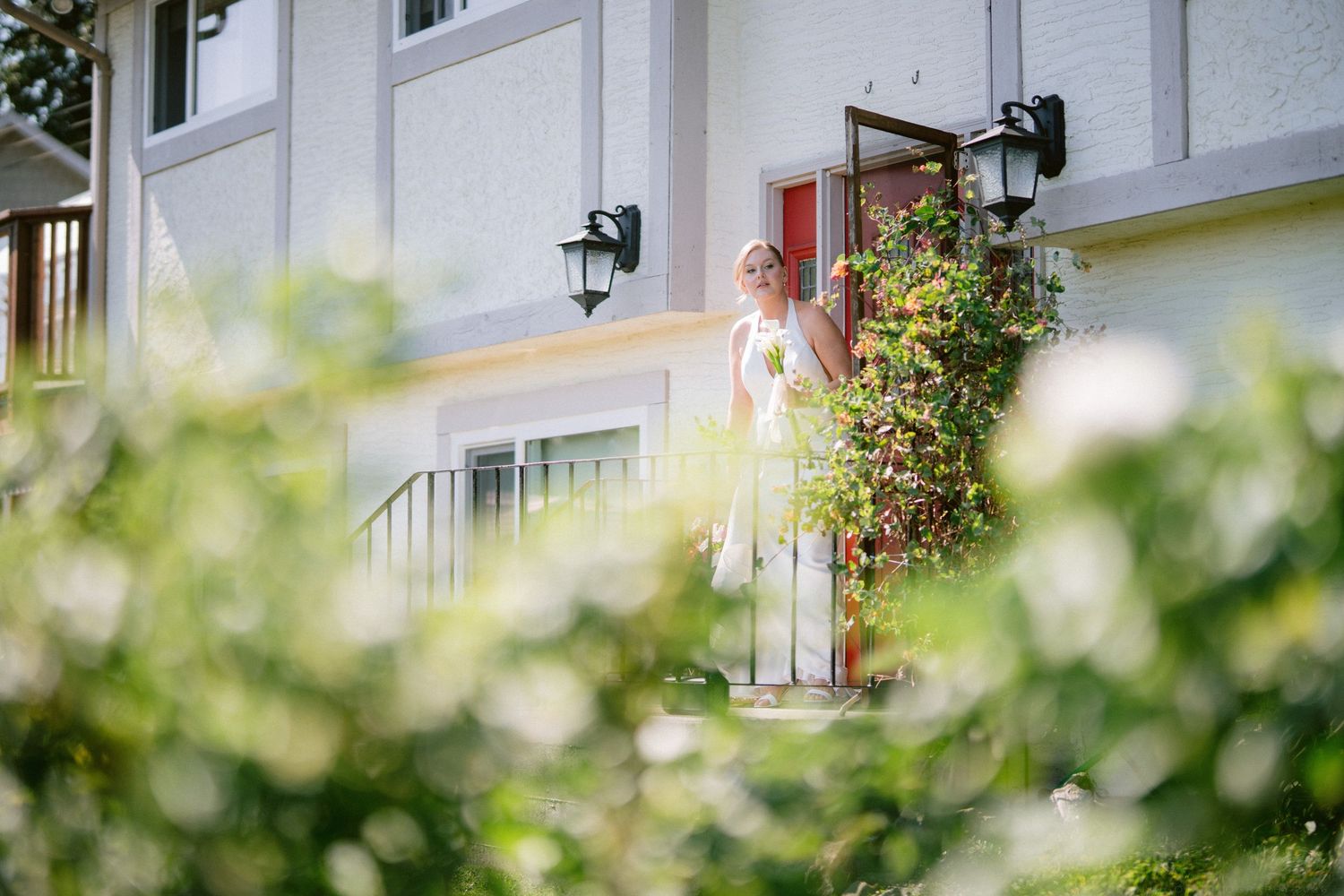 Bride in white dress standing at the entrance of a home surrounded by lush green bushes and white flowers.
