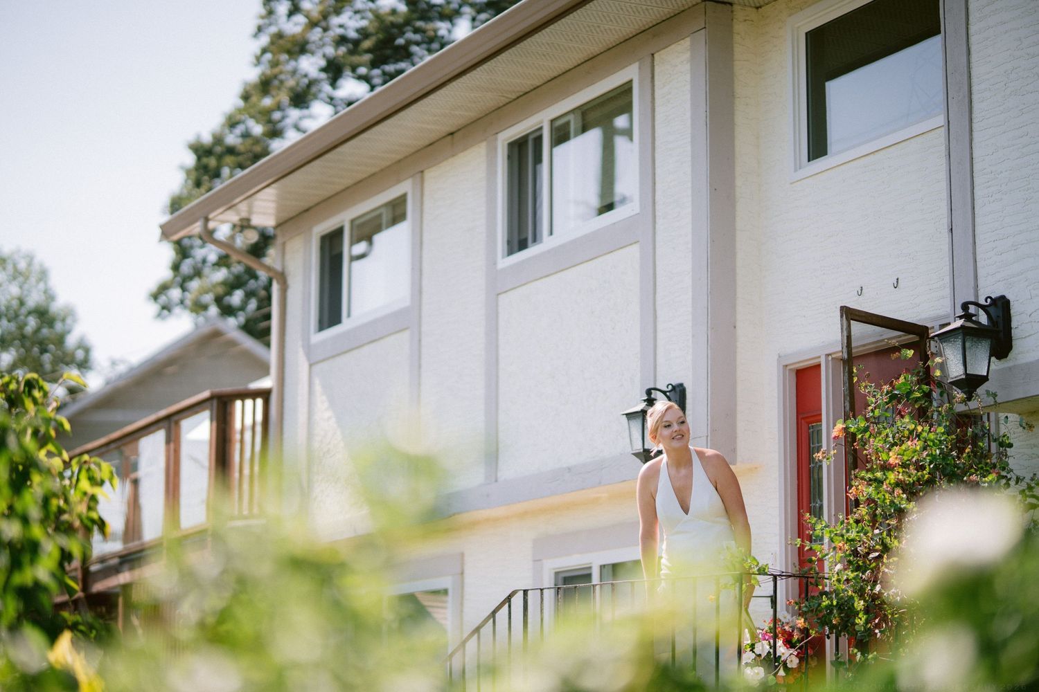 Bride in white wedding dress standing on steps outside a charming white house surrounded by lush greenery.