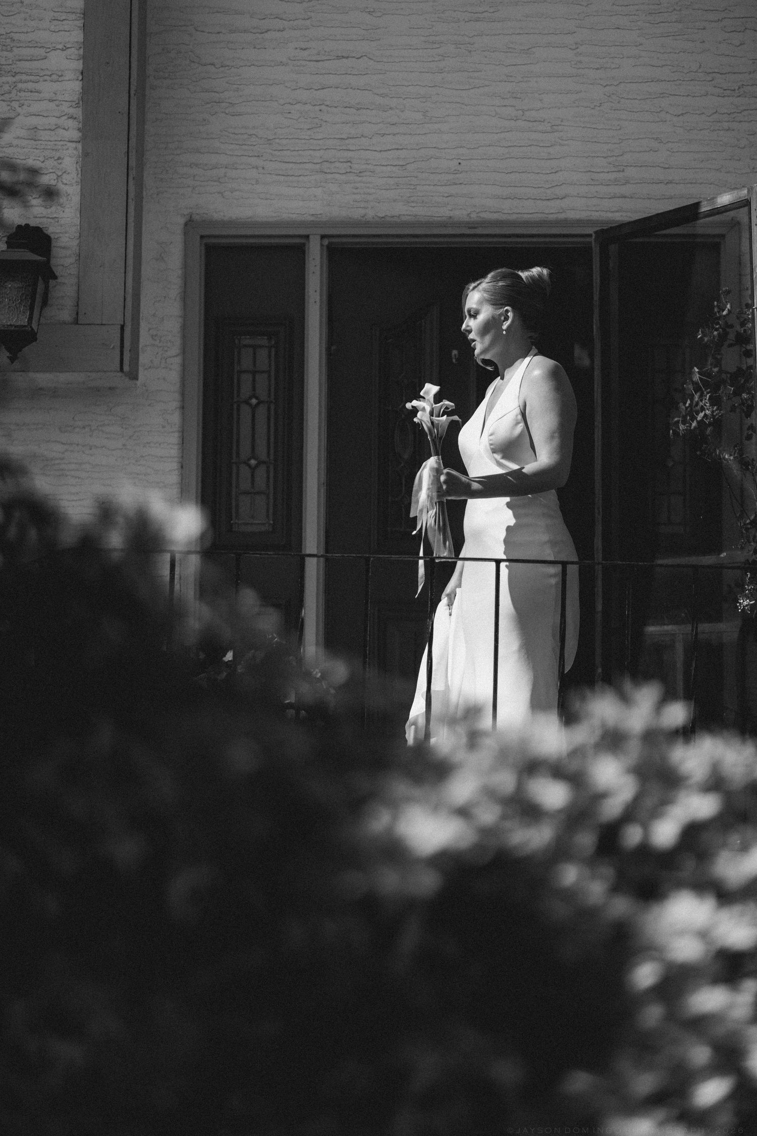 Elegant bride in white halter dress holding flowers standing on porch steps in black and white photo.