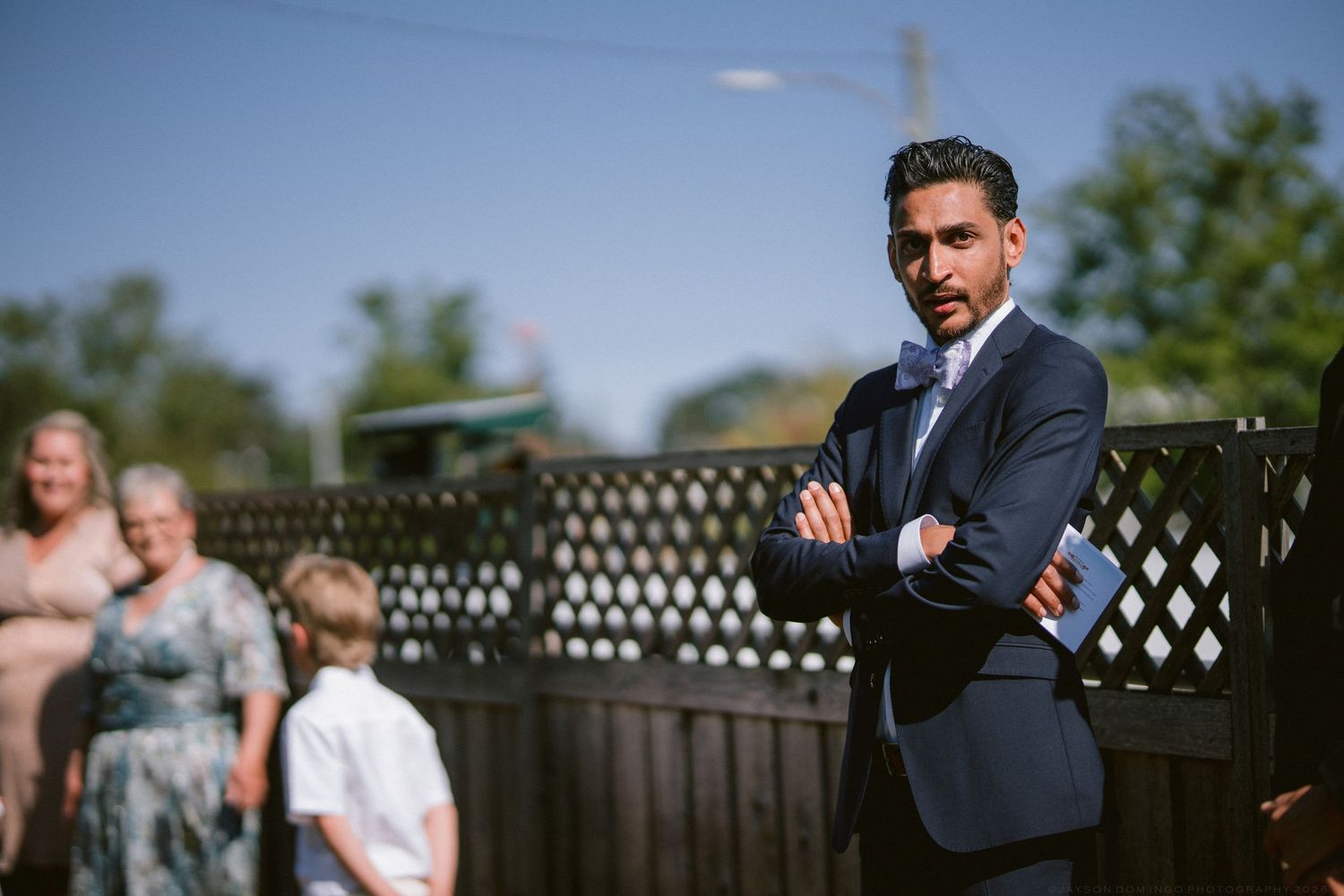 Groom in navy suit standing with arms crossed at outdoor wedding ceremony with guests in background.