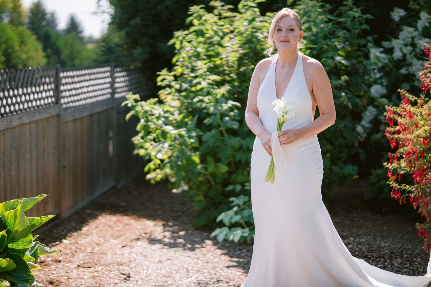 Elegant bride in a white halter gown holds a calla lily bouquet while standing in a lush garden pathway.