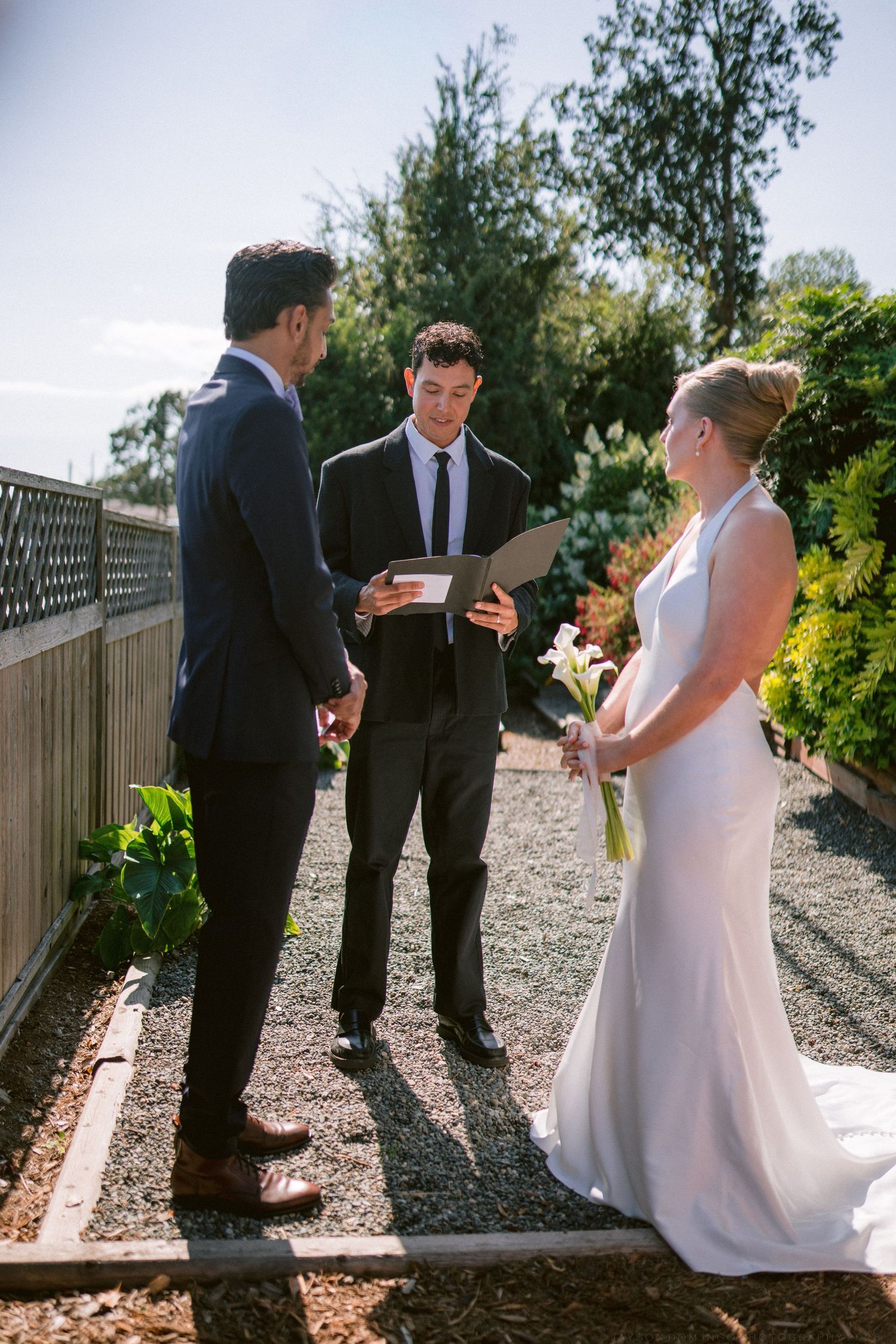 Outdoor garden wedding ceremony with bride, groom, and officiant exchanging vows in sunlight.