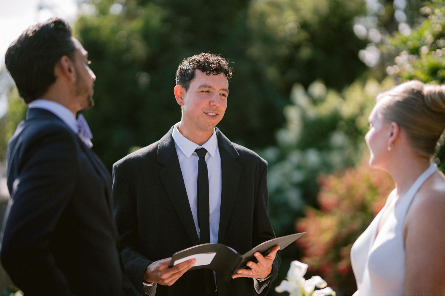 Officiant in black suit reading vows to interracial couple during intimate outdoor wedding ceremony.