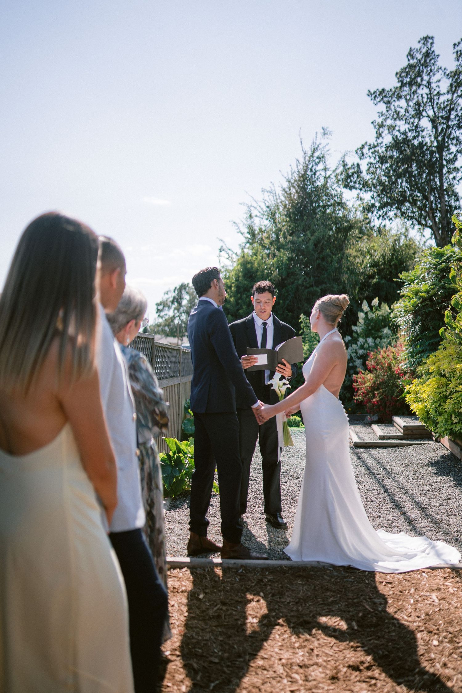 Outdoor wedding ceremony with bride and groom holding hands before an officiant in a sunlit garden setting.