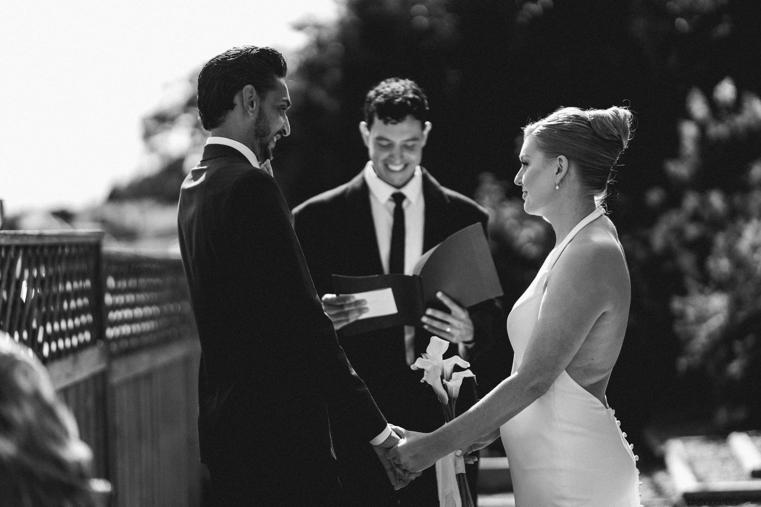 Bride and groom exchanging vows with smiling officiant in black and white ceremony photo.