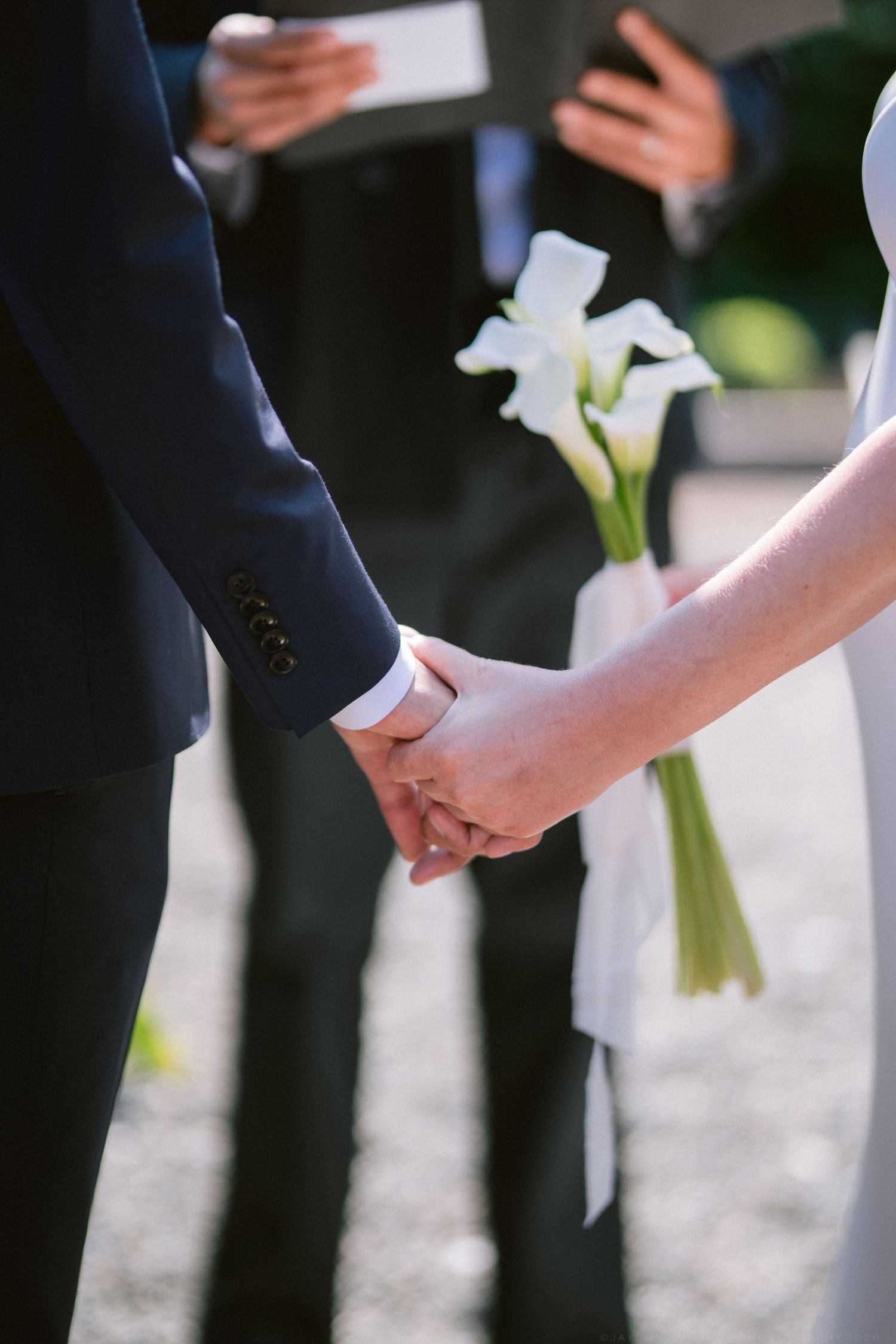 Close-up of bride and groom holding hands during wedding ceremony, bride clutching white calla lily bouquet.