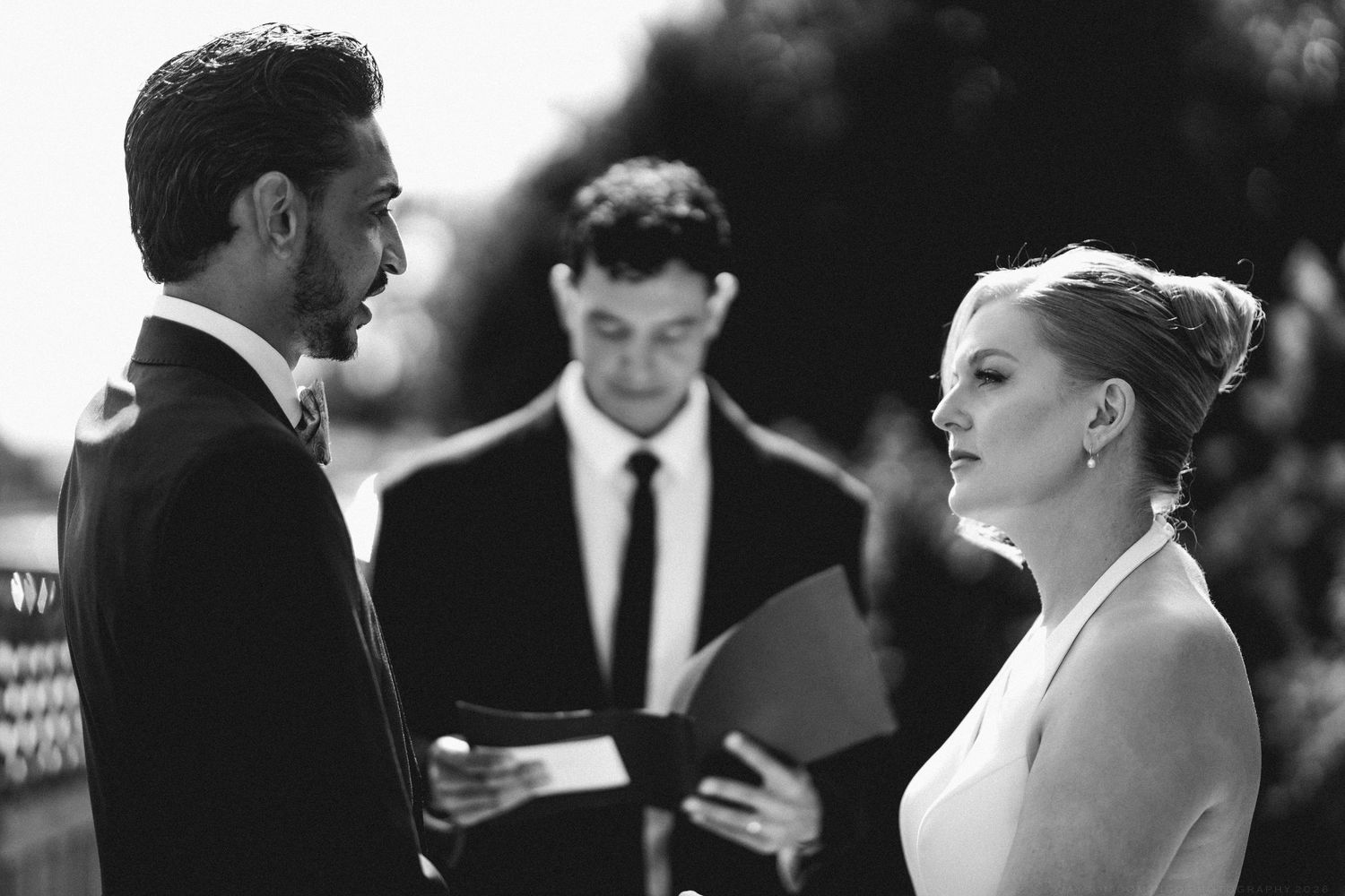 Black and white photo of couple exchanging vows with officiant during outdoor wedding ceremony.