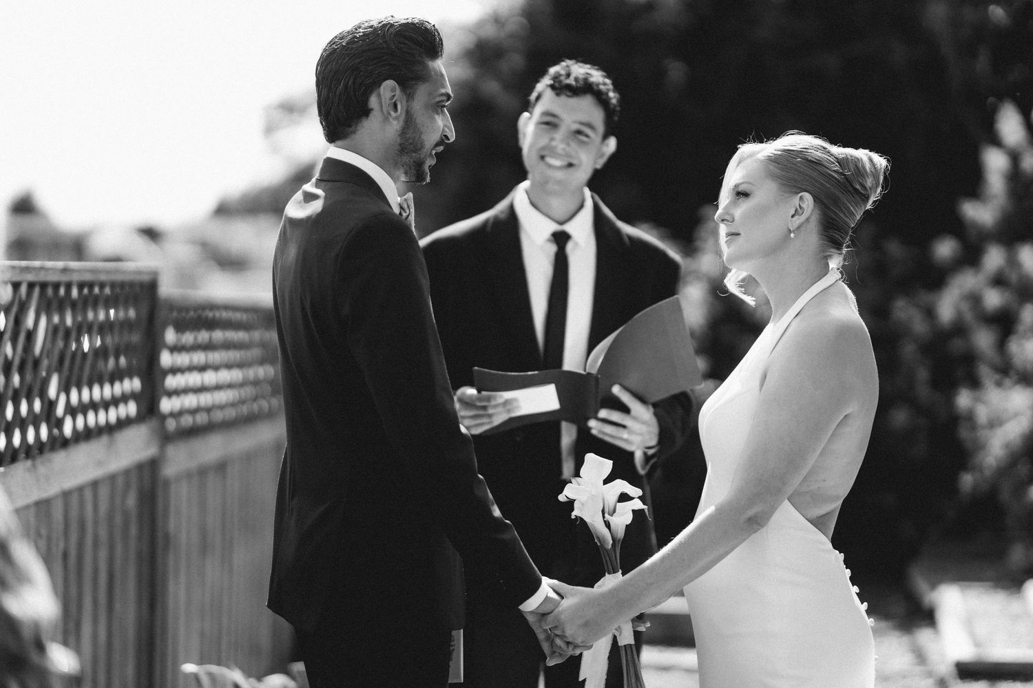 Black and white photo of smiling bride and groom exchanging vows with officiant during outdoor wedding ceremony.