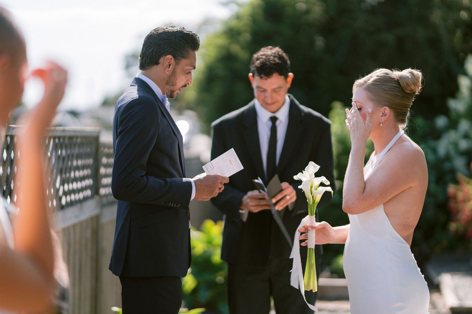 Groom reading vows while emotional bride holds calla lilies during outdoor wedding ceremony.