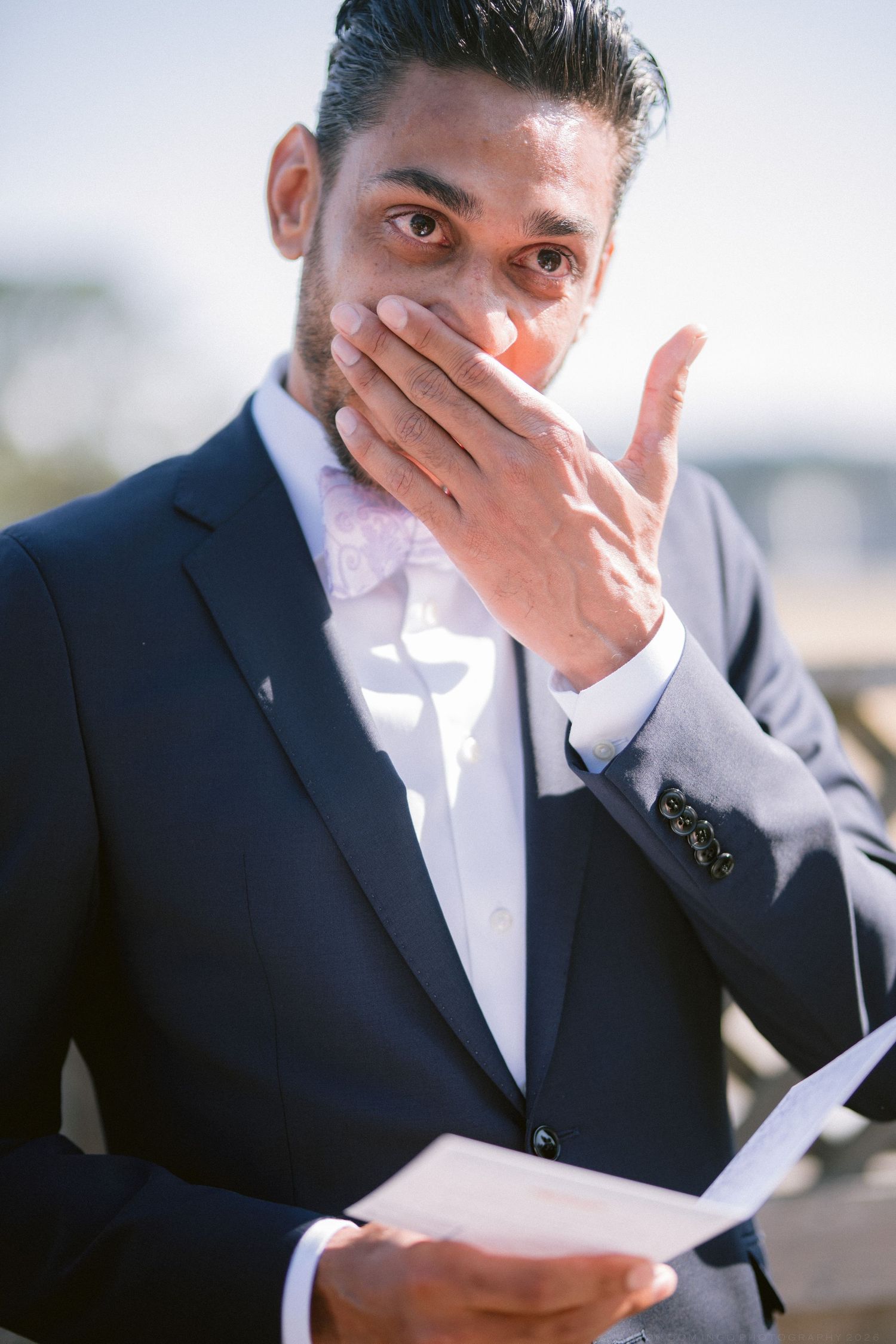 Emotional groom in navy suit covering his mouth while reading wedding vows outdoors in bright sunlight.