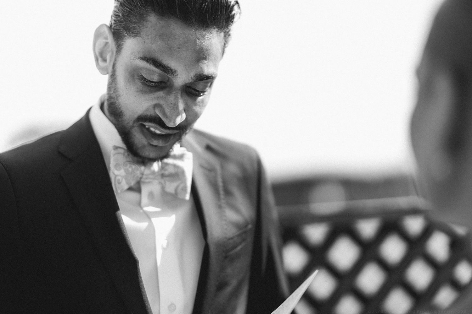 Emotional groom in black tuxedo reading wedding vows in intimate black and white close-up photo.