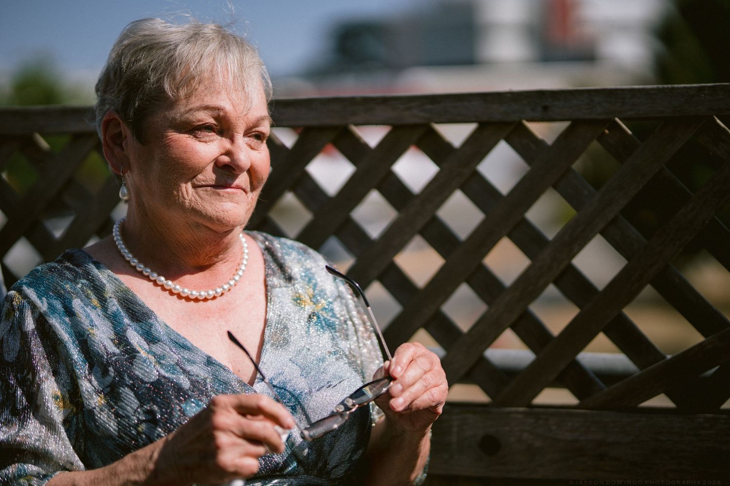 Elegant senior woman with pearl necklace smiles warmly while seated on a wooden bench at a wedding.