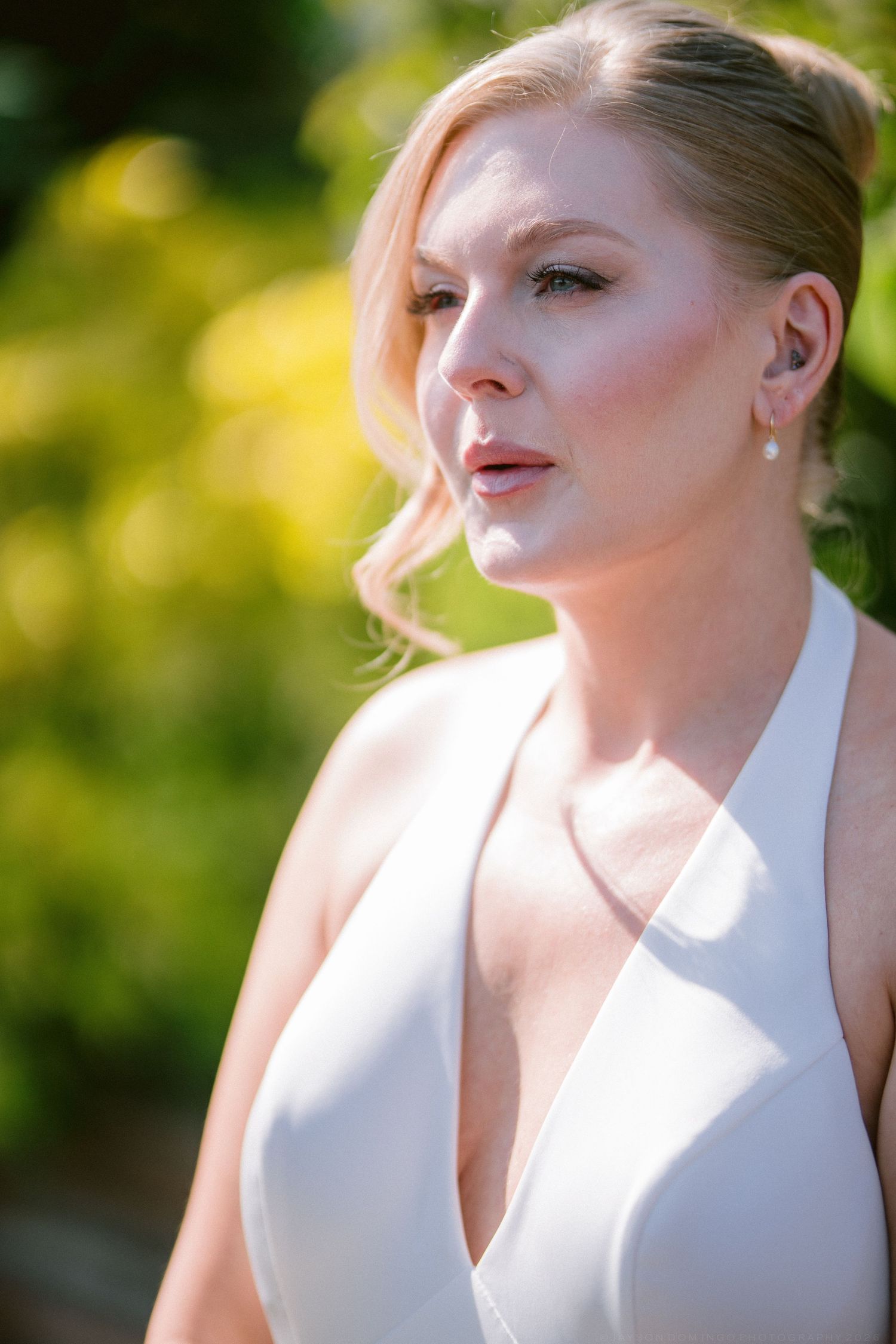Beautiful blonde bride in white halter wedding dress with soft bokeh green garden background.