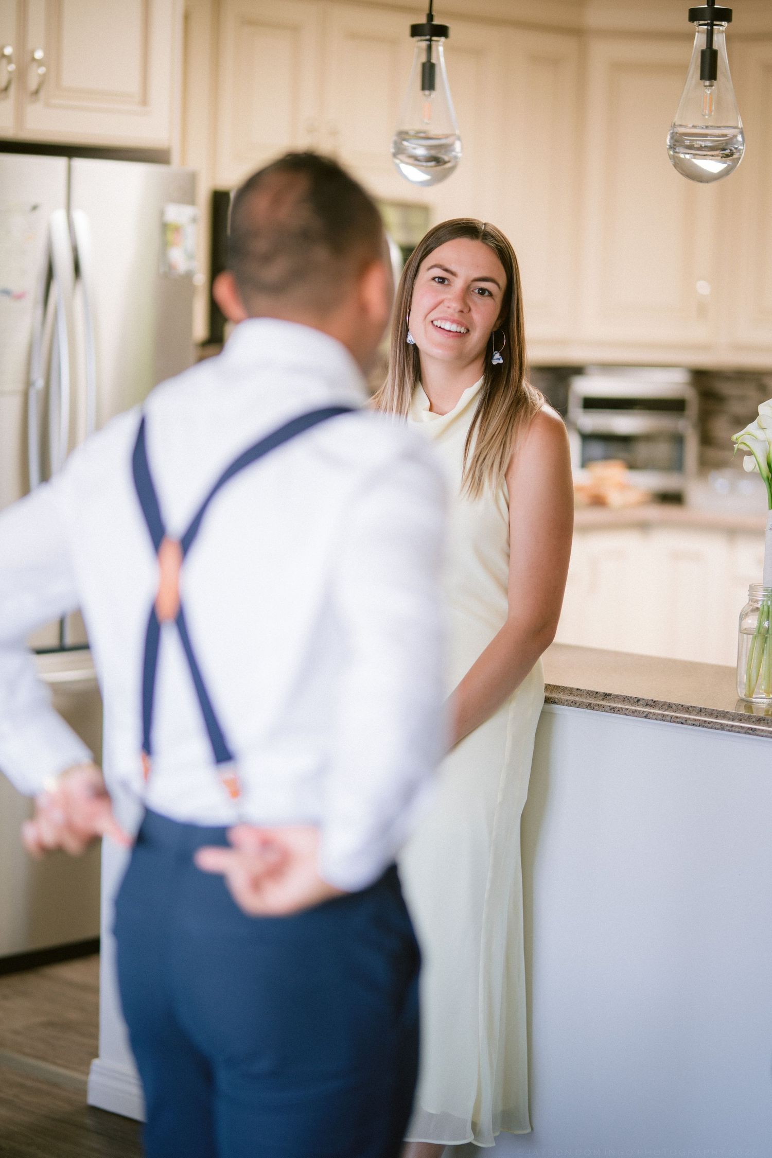 Smiling bridesmaid in sage green dress greeting groomsman in white shirt with navy suspenders in kitchen.