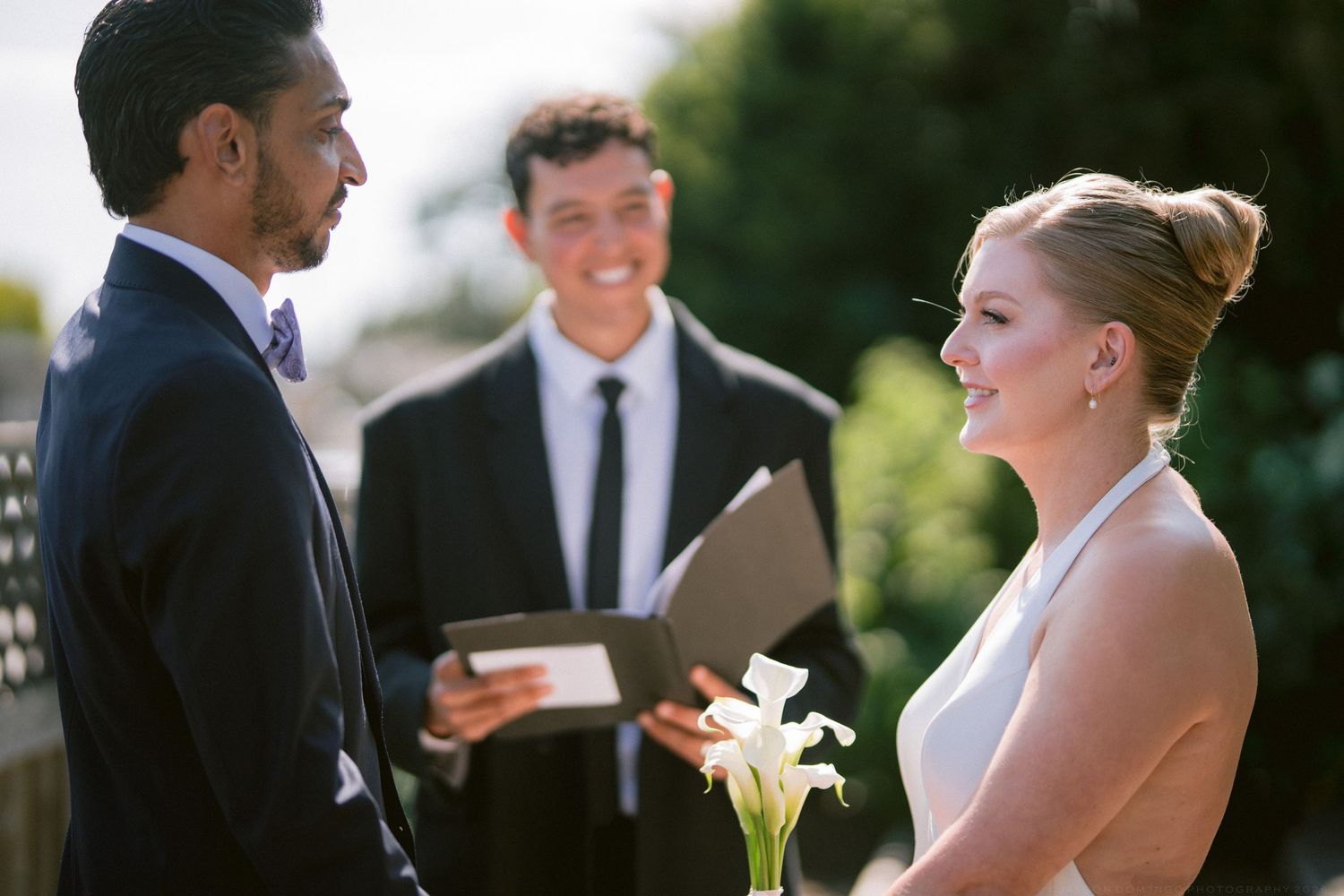 Bride and groom exchanging vows outdoors with smiling officiant holding ceremony book behind them.