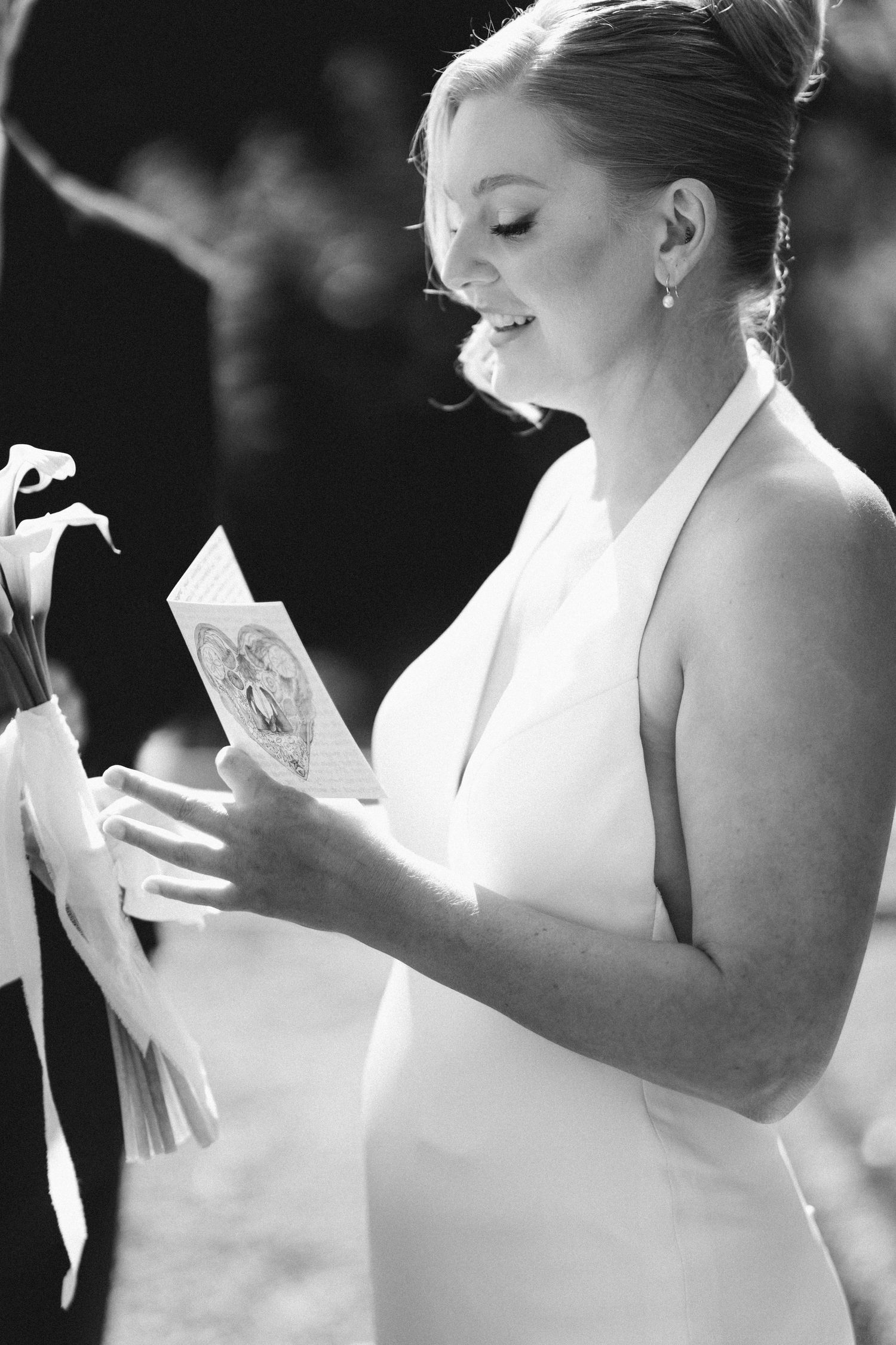 Emotional bride in white halter dress reading vows card while holding calla lily bouquet, black and white.