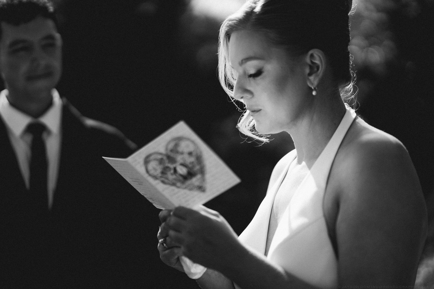 Bride reading heartfelt vows from illustrated card during outdoor wedding ceremony in black and white.