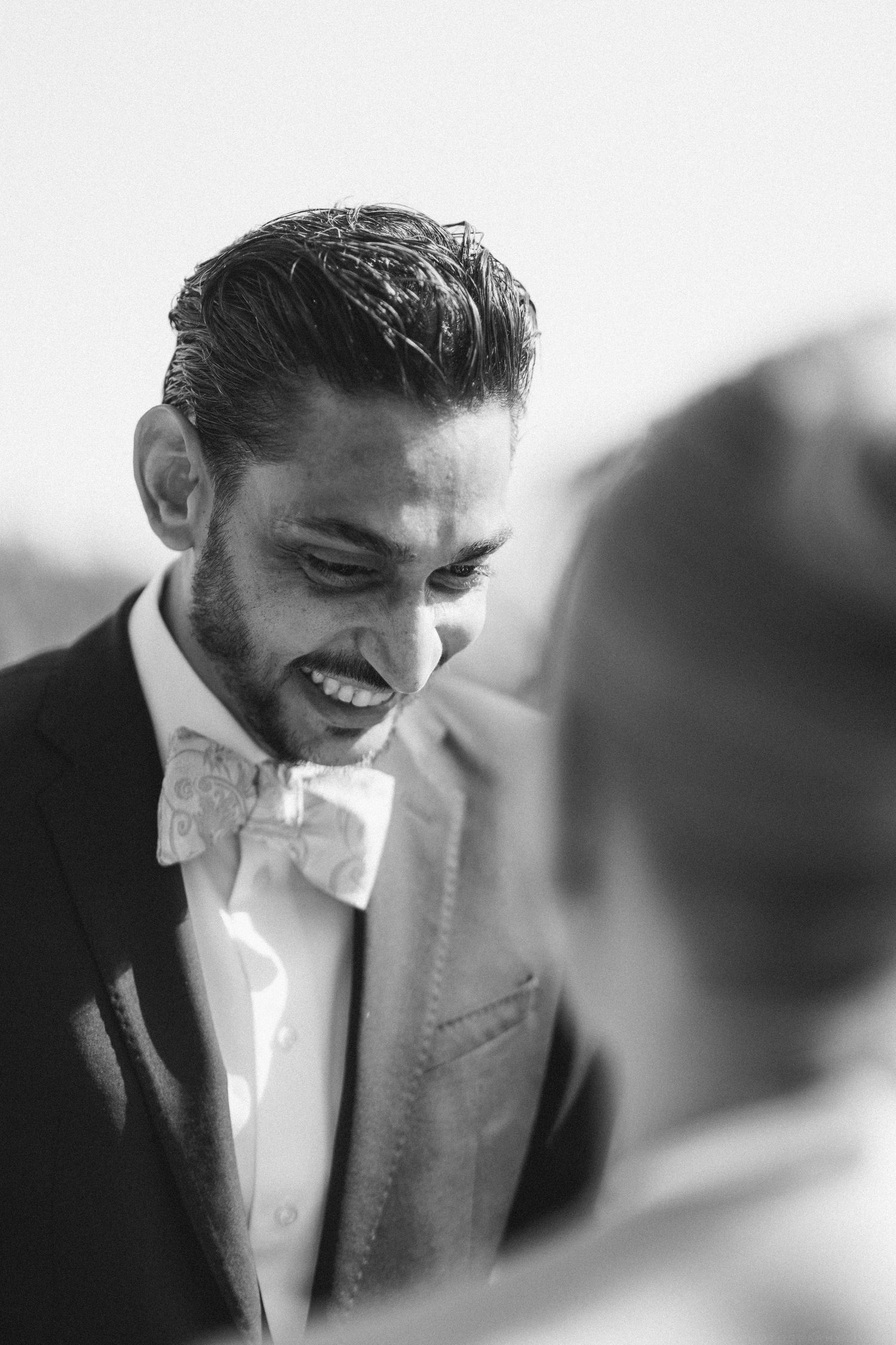Smiling groom in suit and floral bow tie at wedding ceremony, black and white portrait photography.