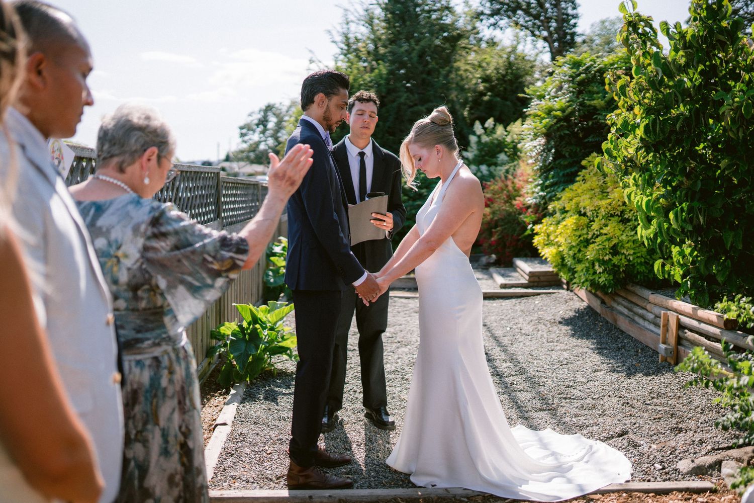 Outdoor wedding ceremony with couple holding hands while officiant and guests gather in a garden setting.