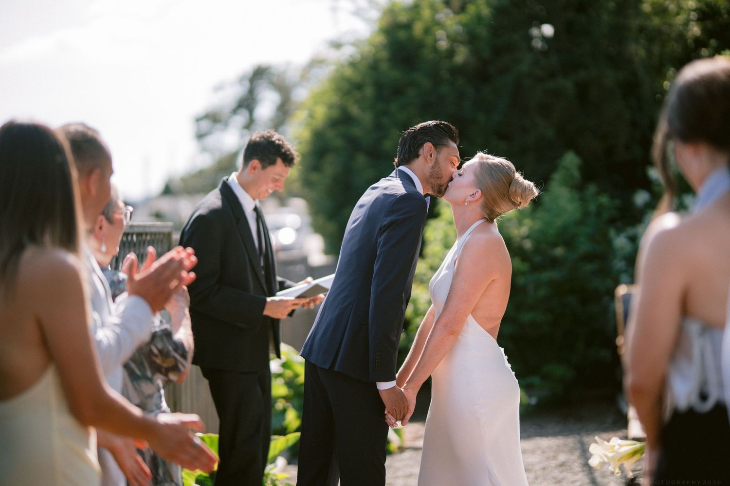 Bride and groom share first kiss while guests applaud at sunny outdoor wedding ceremony.