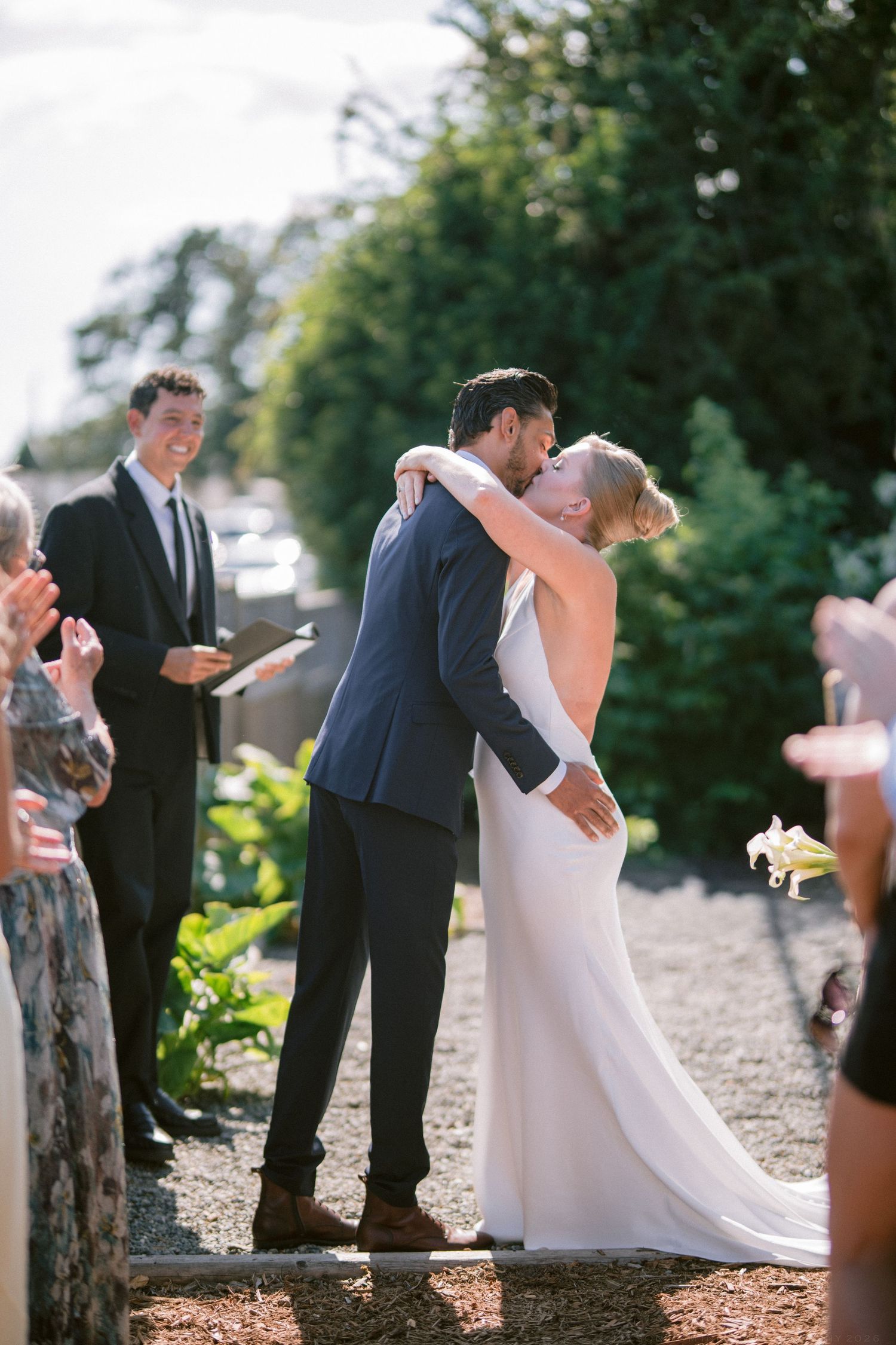 Newlyweds share their first kiss as married couple while guests applaud at outdoor garden wedding ceremony.