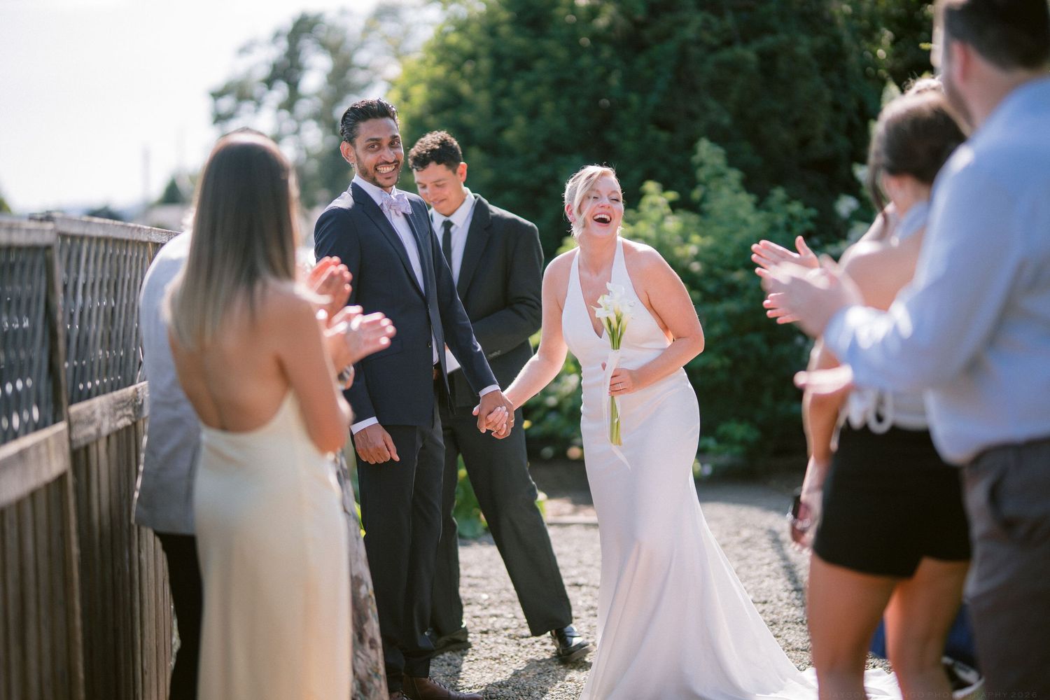 Joyful newlyweds walking hand in hand as guests applaud and celebrate after outdoor wedding ceremony.