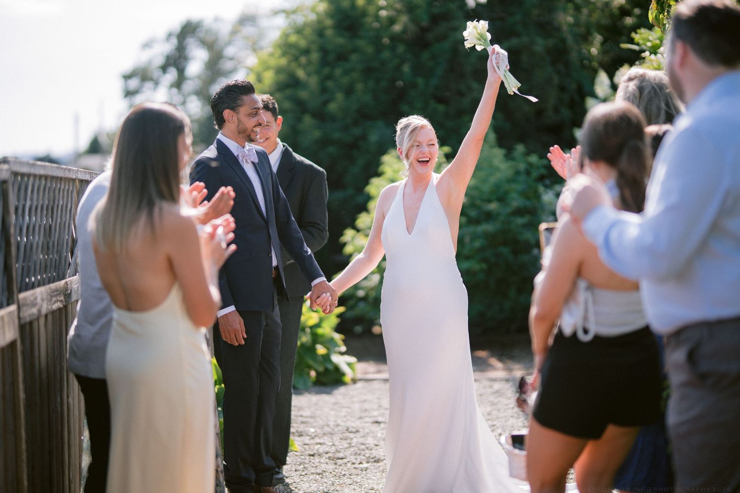 Joyful bride raising bouquet while walking with groom through cheering wedding guests outdoors.
